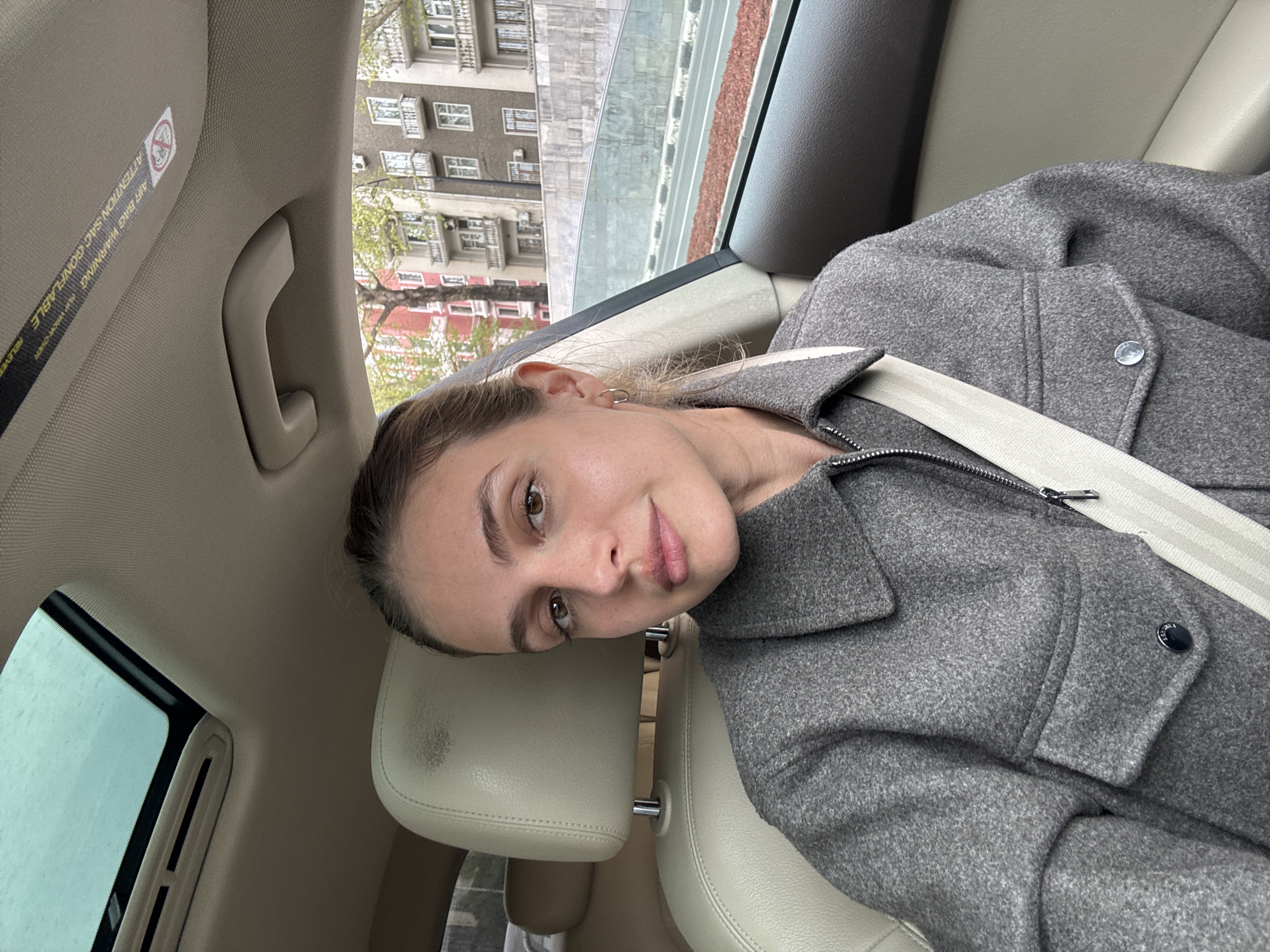 A young woman is depicted in a close-up, selfie-style shot, seated in the passenger seat of a car. Her head is gently tilted and rests against a light-colored, possibly leather, headrest. She looks towards the camera with a calm expression, featuring a subtle, soft smile. She has brown hair and appears to be wearing light makeup.
The woman is dressed in a grey, textured jacket or coat, and a light-colored seatbelt is fastened securely across her chest and shoulder. The car's interior is predominantly light-colored (beige or tan), including the seats and the overhead lining. A grab handle is visible on the ceiling.
Through the car window, an urban street scene is visible. Multi-story buildings with numerous windows and balconies line the street, some with a darker, grayer facade and others with hints of reddish tones. Trees, some with sparse foliage, are also present, suggesting a temperate season. The lighting suggests it is daytime, with a soft, diffused light, possibly indicating an overcast sky.
A warning label is visible on the car's sun visor or overhead panel, clearly showing "AIR BAG WARNING" and "ATTENTION SAC GONFLABLE WARNING" text. The scene captures a relaxed moment within a vehicle in an urban environment, identified as Tbilisi, Georgia.