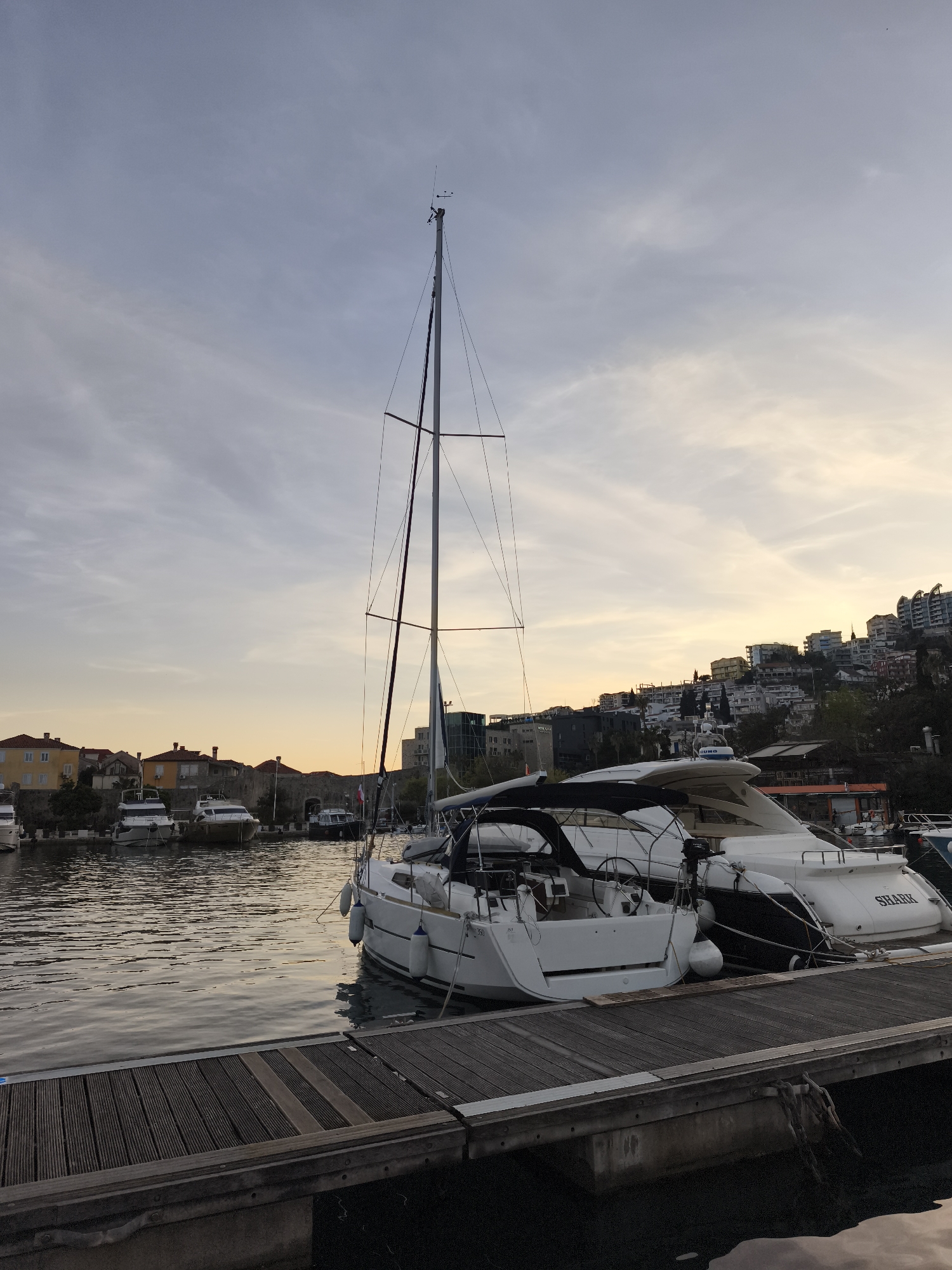 A medium-angle outdoor shot shows several boats moored to a wooden dock in a harbor, with buildings on hillsides in the background under a cloudy sky.
The primary subject is a white sailboat with its mast towering upwards, docked on the right side of the frame. To its right is a larger, white motor yacht with the name "SHARK" visible on its stern. The wooden dock is composed of planks and extends from the bottom of the frame towards the boats.
In the background, across the water, are various buildings, including residential structures and a modern glass-fronted building. Further back, the hillsides are dotted with more buildings and vegetation. The sky is overcast, with hints of orange and yellow near the horizon, suggesting either sunrise or sunset. The water is calm, with reflections of the boats and buildings visible.