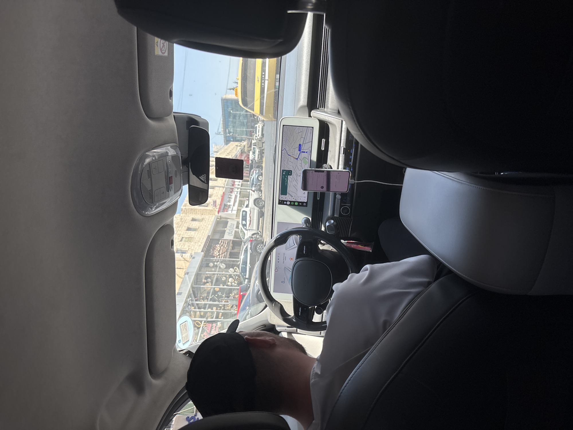 A passenger's point-of-view shot taken from inside a modern car, looking towards the driver and the road ahead. The driver, a person wearing a black baseball cap and a white t-shirt, is seated behind the steering wheel of the car. The car's interior is visible, featuring a large digital display for navigation and other vehicle information, as well as a mobile phone mounted on the dashboard. Outside the car, a street scene unfolds with other vehicles, buildings, and shops, suggesting an urban environment. The yellow taxi in the background identifies the car as a taxi. The overall scene depicts a typical journey within the city of Dabouq, Jordan.