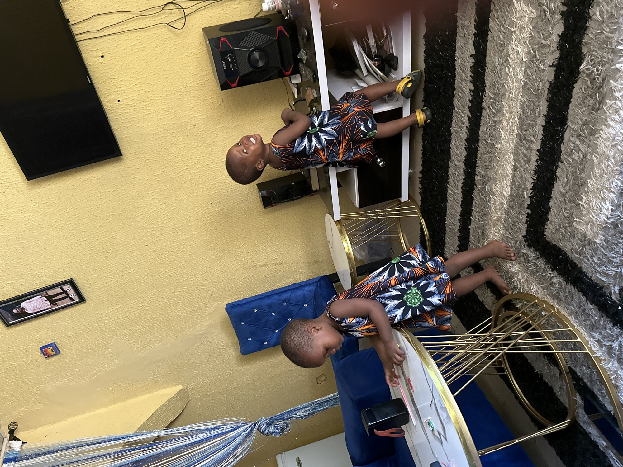 In a living room located in Narayi, Nigeria, two young children are captured in a candid moment. The room is decorated with yellow walls, a large black television mounted on the wall, and a blue velvet chair with diamond-like studs. The floor is covered with a shag rug featuring black and gray stripes.
In the foreground, a young girl with short hair is reaching out towards a white marble coffee table with a gold metal frame. She is wearing a colorful sleeveless dress with a floral print. On the table, there are a few small objects, including a black rectangular item with a red strap, and what appear to be some colorful sticks.
In the background, another child, also a young girl, is playfully looking towards the camera and smiling. She is standing near a black speaker system on a shelf unit. She is wearing a similar colorful sleeveless dress and yellow footwear. The environment suggests a home setting, with elements of decor and entertainment visible.