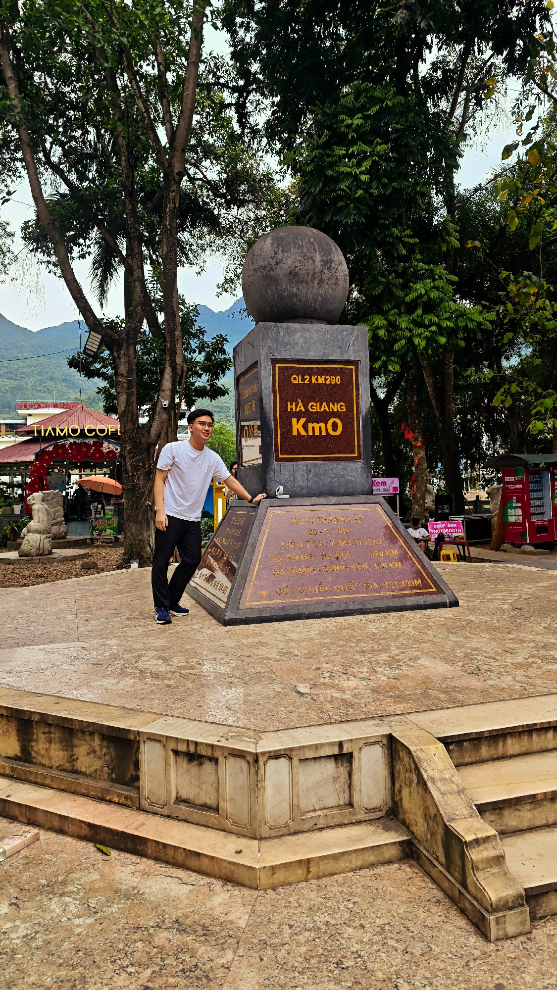 In Hà Giang 1 Ward, Vietnam, a young man in a white t-shirt and black pants stands near a monument, casually touching its base with his right hand. The monument, which is a gray rectangular structure topped with a gray sphere, has signs that read "QL2 KM290 HÀ GIANG Kmo". The monument also contains an inscribed plate. The man is smiling and wearing glasses.
Behind the monument are trees and a glimpse of mountains in the distance. To the left, a building with a sign that reads "TIAMO COFI" is visible. There is a statue and red decorations near this building. To the right, there is a vending machine. The monument is on a raised platform with steps leading up to it. The plaza where the monument is located appears well-worn.
