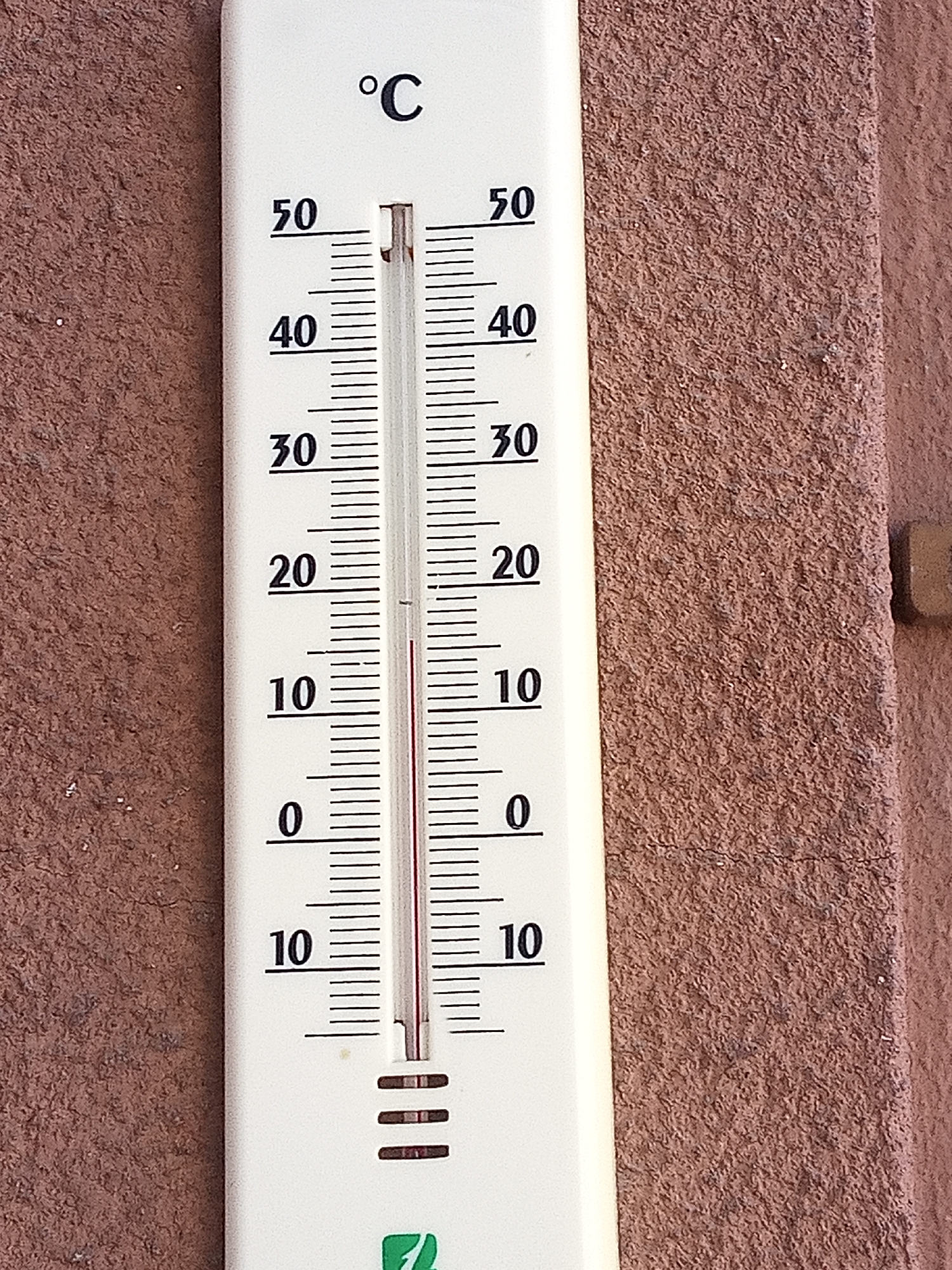 The image shows a white thermometer against a textured reddish-brown wall. The thermometer is marked with degree Celsius markings from 0 to 50, with major increments at 0, 10, 20, 30, 40, and 50. A red liquid column within the thermometer indicates a temperature between 25 and 30 degrees Celsius. The thermometer is mounted on a wall in what appears to be an outdoor setting. No people or actions are visible. The environment is characterized by the rough texture of the wall. The exact location within Sant Joan Les Fonts, Spain, is not discernible from the visual information provided in the image.