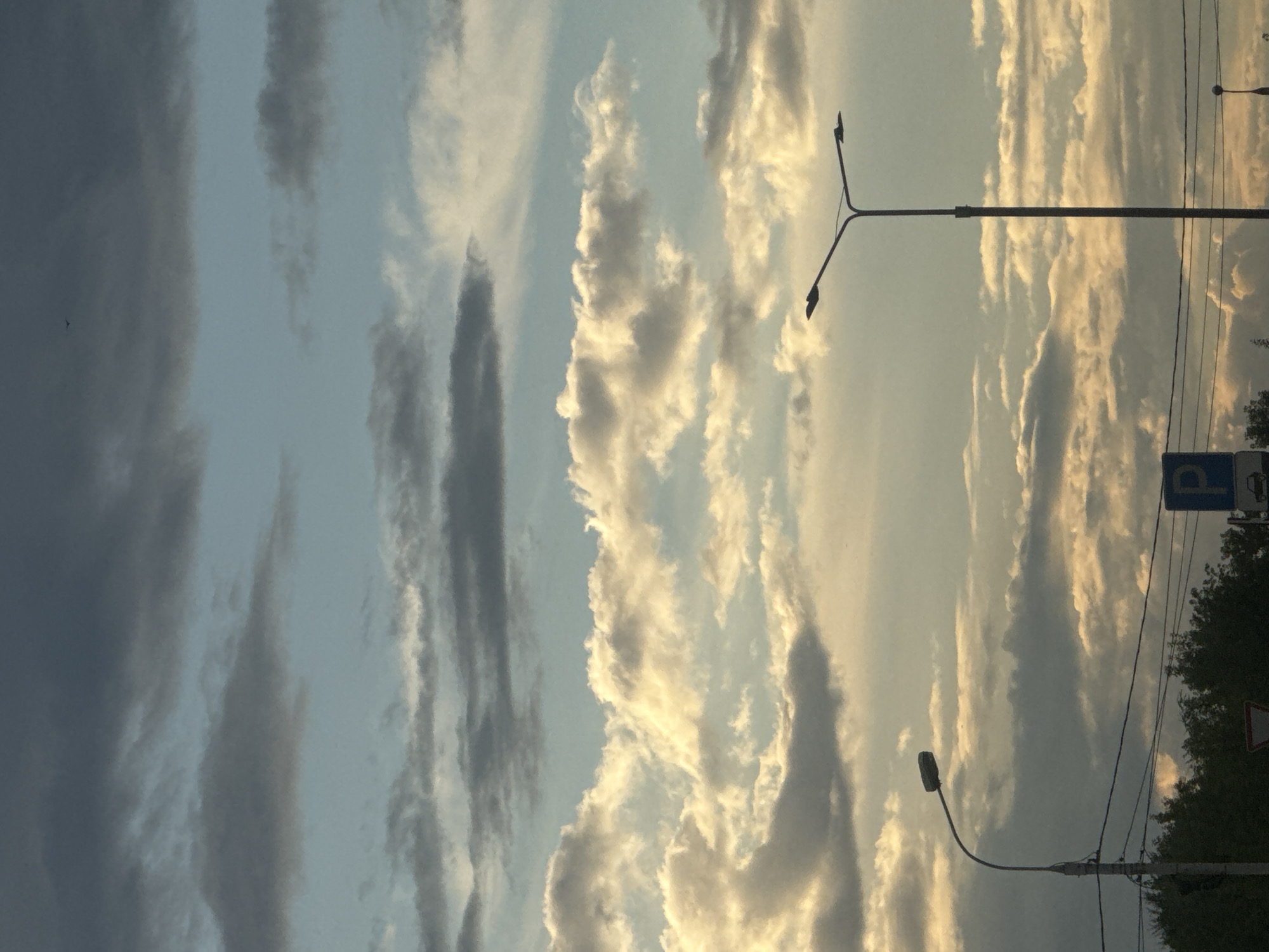 The image captures a sky filled with a mix of dark and light clouds, with a hint of blue sky peeking through. Two streetlights are visible, one on the left with a single lamp and another on the right with two lamps. A blue parking sign with a white 'P' is also present, along with some trees and power lines in the background. The lighting suggests it might be late afternoon or early evening. No specific landmarks identifying Irkutsk, Russia, are apparent in this generic urban or suburban scene.