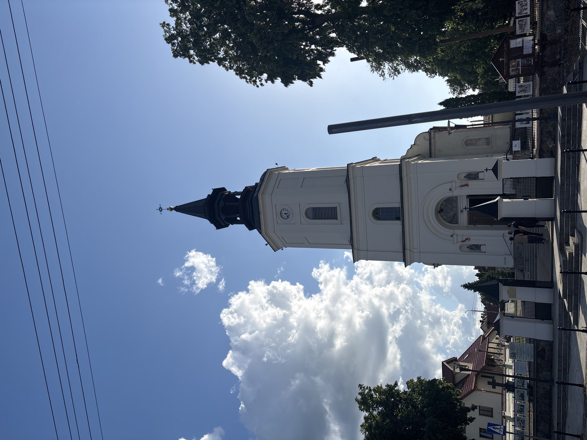 The image captures a frontal view of a white church with a tall, black-spired bell tower, situated in Sobolewo, Poland. The church's facade features a large arched entrance and a clock on the tower. In front of the church, several people are visible near the entrance. The church is framed by lush green trees on the right and left sides. In the background, a residential building with a red roof and a clear blue sky with scattered white clouds are visible. The scene appears to be a sunny day.
