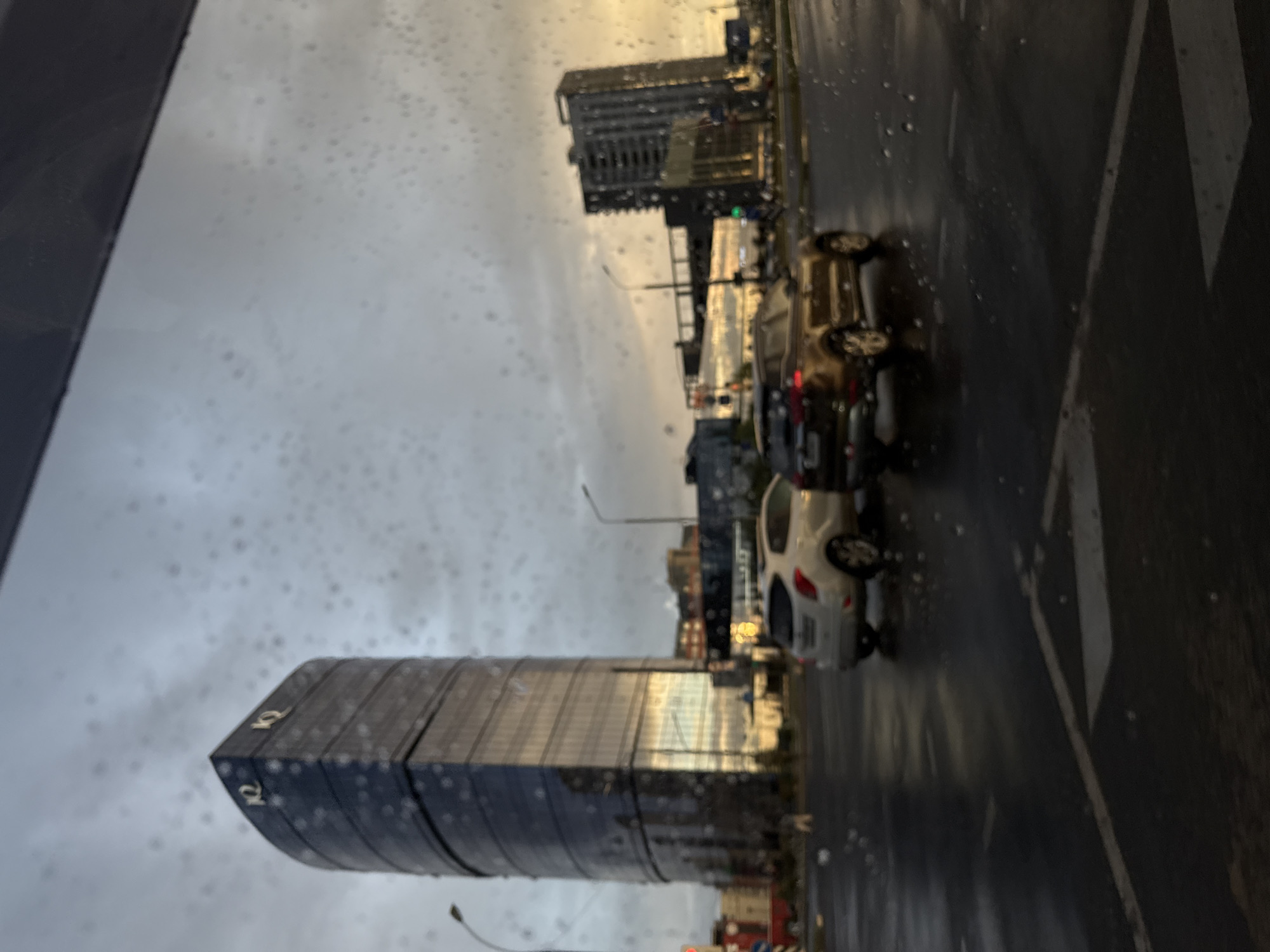 This is a slightly blurry, low-angle, eye-level shot of a city street in Brest, Belarus, taken through a window with raindrops on it. A distinctive curved, glass-facade skyscraper with "IQ" logos is prominently featured on the left side of the frame. In the foreground, a wet asphalt road with lane markings is visible, with two cars driving on it. The car on the left is a white hatchback, and the car on the right is a brown SUV. The sky is overcast with a warm glow of sunset or sunrise visible in the distance on the right. Another modern, rectangular building is visible behind the brown SUV. There are no people clearly visible in the frame. The overall impression is an urban setting on a rainy day.
