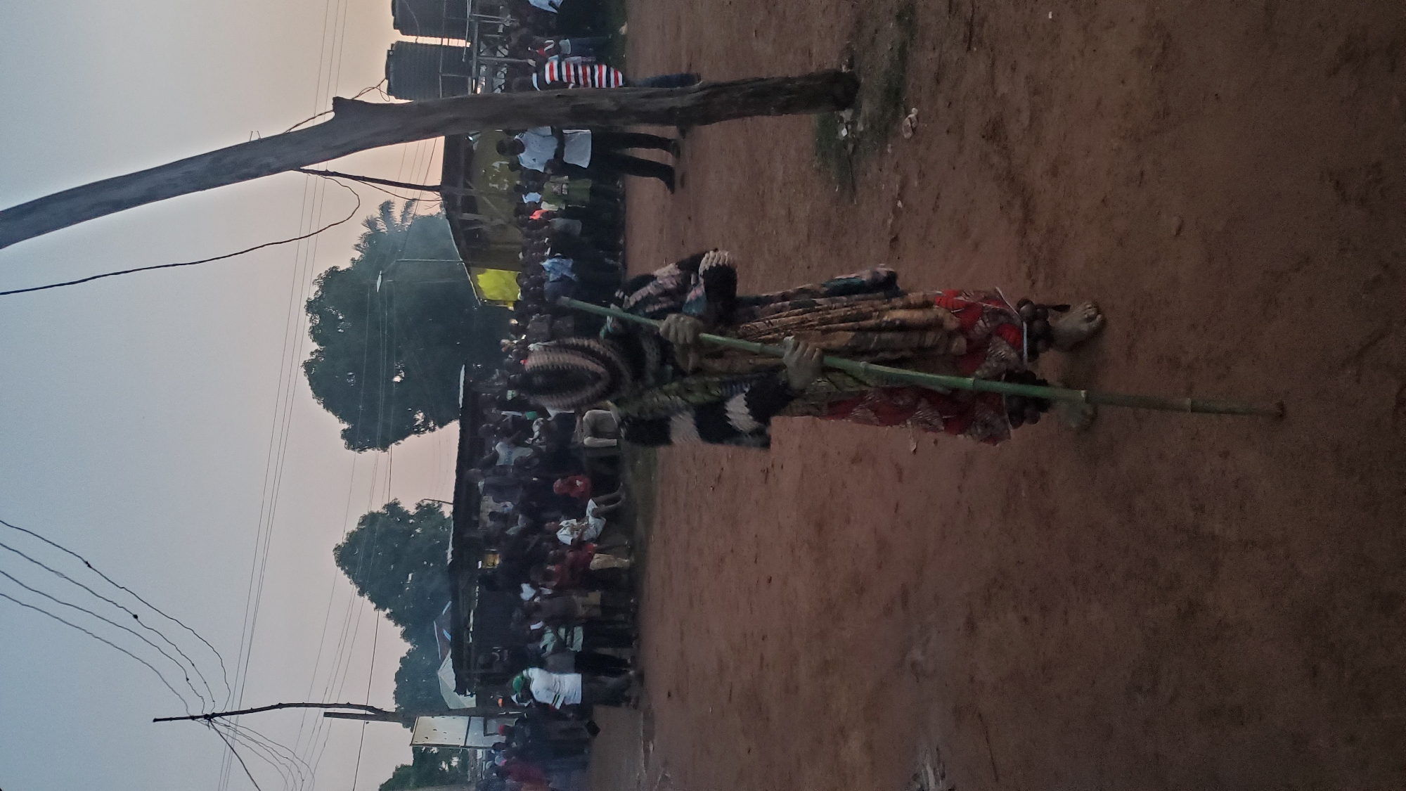A person in a colorful, patterned costume and mask stands in a dirt clearing, holding a long green stick. They are facing forward, with their feet bare and adorned with dark beads around their ankles. In the background, a crowd of people gathers, some standing and others seated. Behind the crowd, there are wooden structures, trees, and utility poles with overhead wires. The sky is overcast and suggests either dawn or dusk. The scene appears to be set in Akwanga, Nigeria, based on the provided context.