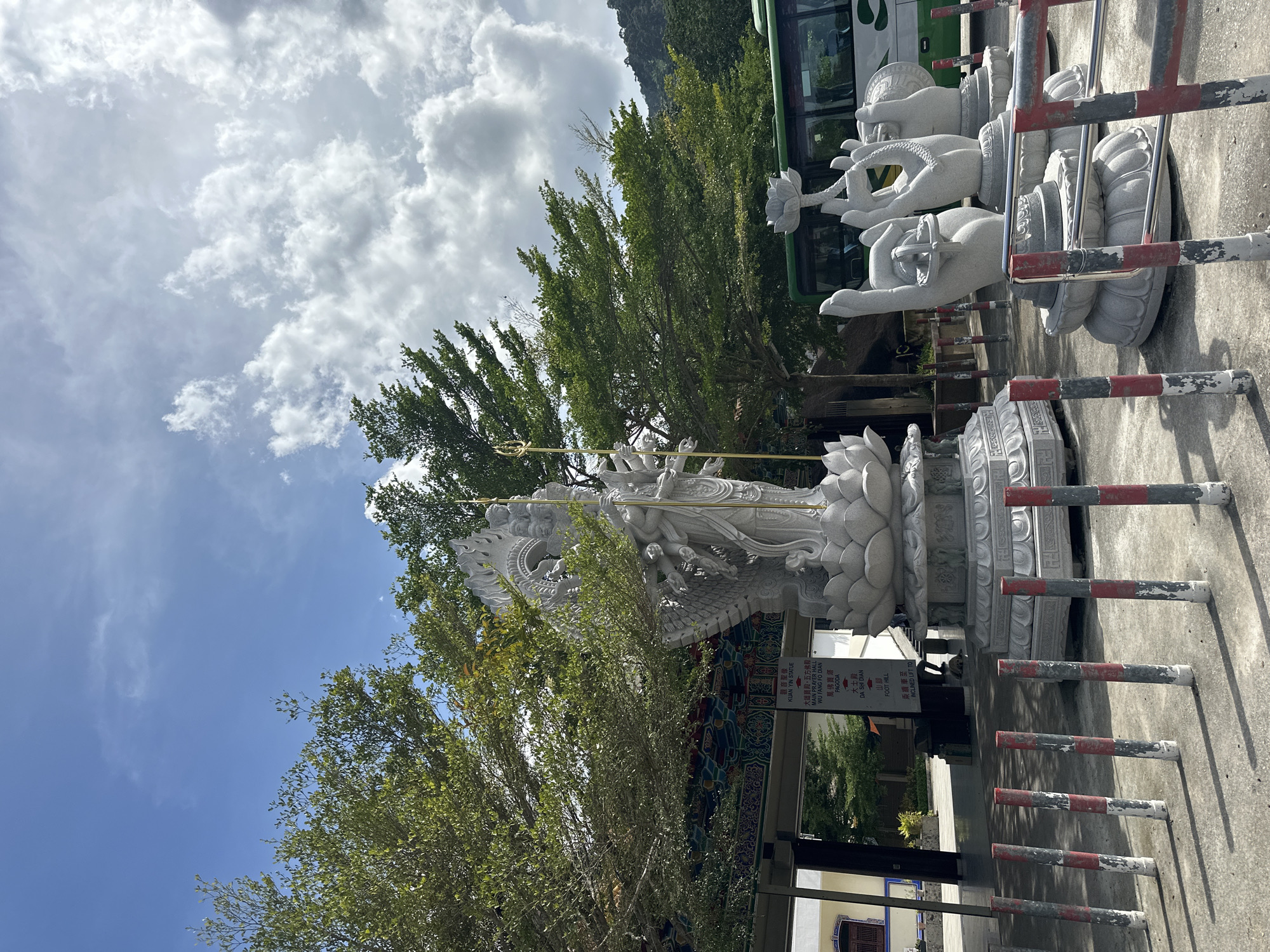 In George Town, Malaysia, this image captures a bright, sunny day with a partly cloudy sky and lush green trees. In the foreground, several cylindrical bollards with red and white markings line a concrete path, separating it from other areas. To the right, multiple stone statues of hands, each holding a lotus flower, are arranged on a circular base. Further back, a large, intricately carved white stone statue of a multi-armed deity stands on a tiered lotus-shaped pedestal. Behind the deity, a colorful, traditional architectural overhang is visible, adorned with intricate patterns. A green bus is parked behind the statues, and in the background, a hillside covered in dense vegetation can be seen. Signs with Chinese and English text are mounted on a pole in front of the large deity statue, indicating "KUAN YIN STATUE," "MAIN PRAYER HALL," "PAGODA," and "FOOT HILL."