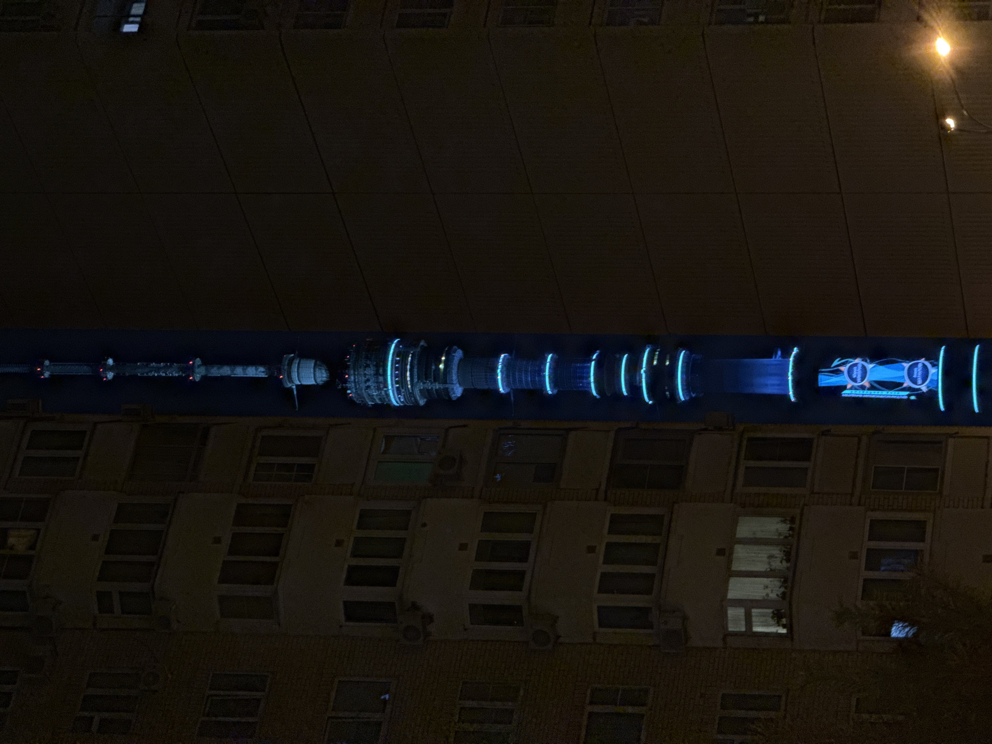 The image depicts a tower with blue lights, possibly a representation of the Moscow Canal, horizontally aligned in the center of the frame. It's positioned between two buildings. The buildings have a repeating pattern of windows and what appear to be air conditioning units beneath some windows. The overall impression is a night scene in an urban environment, likely Khimki, Russia. There are lights visible in the upper right corner, possibly street lights. The image exhibits high contrast due to the stark difference between the bright, illuminated tower and the darker buildings.