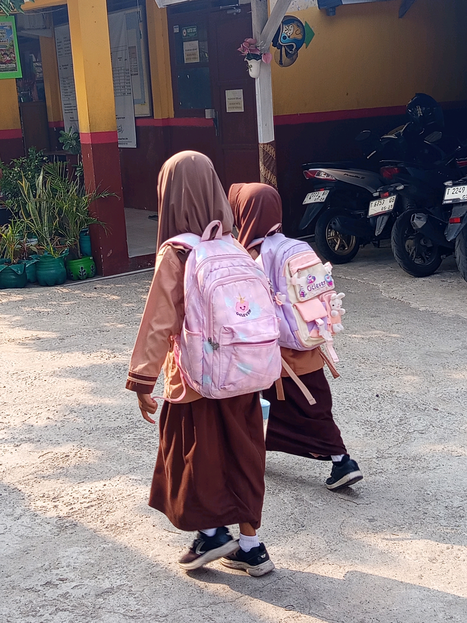 Two young students wearing headscarves and brown skirts walk on a concrete ground in front of a building. They both have backpacks. The student on the left has a pink backpack with a unicorn design. The student on the right has a pink and purple backpack with a bunny charm and the word "Oclever" printed on it. In the background, there are parked motorcycles and a yellow building with red trim. Plants in green pots are also visible to the left. The scene is illuminated by sunlight, casting shadows on the ground. The location is identified as Karawang, Indonesia.