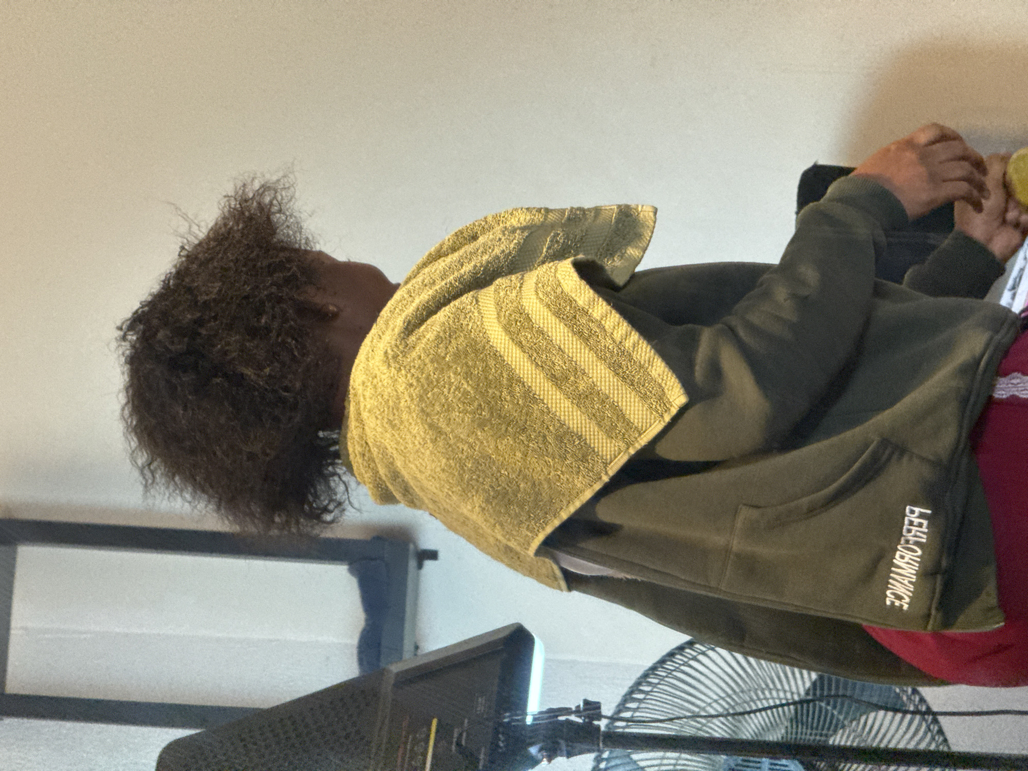 In a domestic setting in Abuja, Nigeria, a person with curly dark hair is partially visible from the back and side. They are wearing a dark green hooded sweatshirt with a white embroidered logo on the left side of the chest, which reads "PERFORMANCE" in reverse. A light green towel with horizontal stripes is draped over their shoulders. The person appears to be holding a yellow object, possibly a fruit like a lemon, in their hands. To the left of the person is a black fan with a wide circular grill and a black speaker on a stand with a bright light attached. A glimpse of a dark framed structure is visible in the background. The lighting suggests an indoor environment.