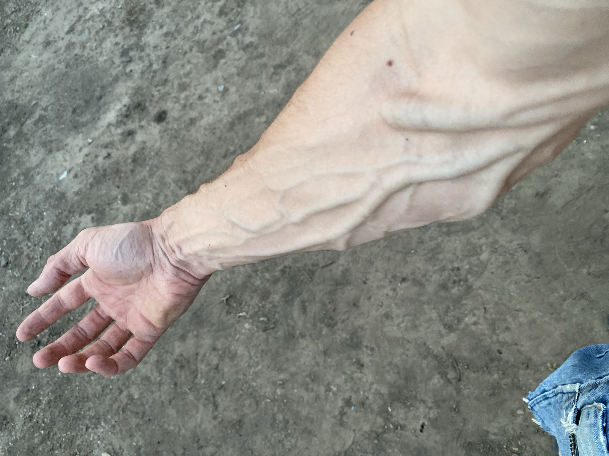This is a close-up, high-angle shot of a person's arm and hand against a grey, granular background. The skin on the arm is pale with prominent blue veins visible due to the lighting and possibly low body fat. The hand is open, fingers slightly curled, and also shows visible veins. The background appears to be a mixture of dirt, small stones, and possibly some debris, suggesting an outdoor, possibly urban or rural, environment. There is a small mole visible on the upper arm. The bottom right corner of the image shows a small portion of blue denim fabric, possibly from clothing. The lighting casts shadows, highlighting the texture of the skin and the ground. The image does not contain any information about the city of Cairaclia, Moldova, or any specific location.
