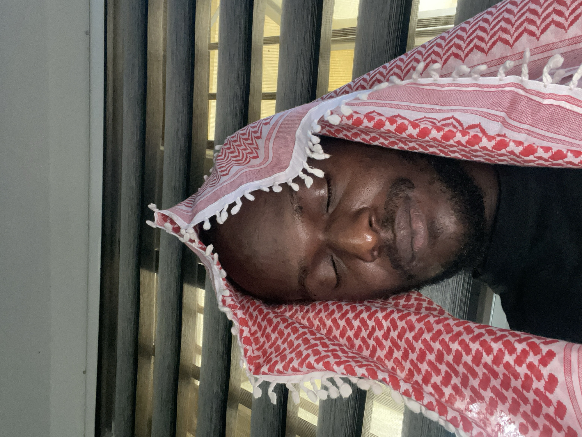 An African male with dark skin is seen from the chest up, wearing a red and white patterned keffiyeh. He has his eyes closed and his head is tilted slightly downwards. He appears to be wearing a black turtleneck. The background consists of horizontal blinds. The lighting suggests it is daytime. The overall scene is a close-up portrait of the man. The location is not discernible from the image itself.