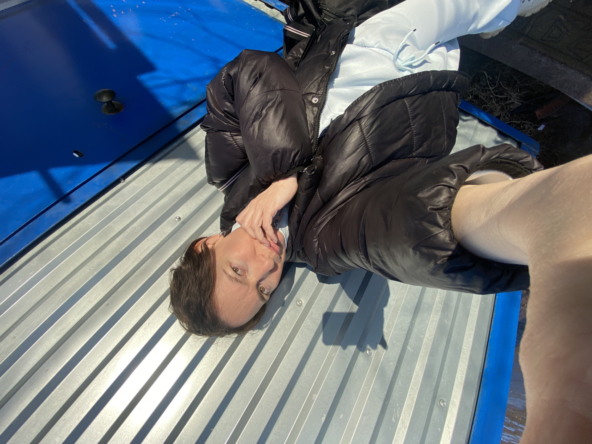 A person is lying on a corrugated metal surface, possibly a roof or a large metal panel. The person is wearing a black puffer jacket and a light blue hooded sweatshirt. A portion of a bright blue structure is visible on the edges of the metal surface. The location and time are not possible to determine, as the city and other important landmarks are not visible.
