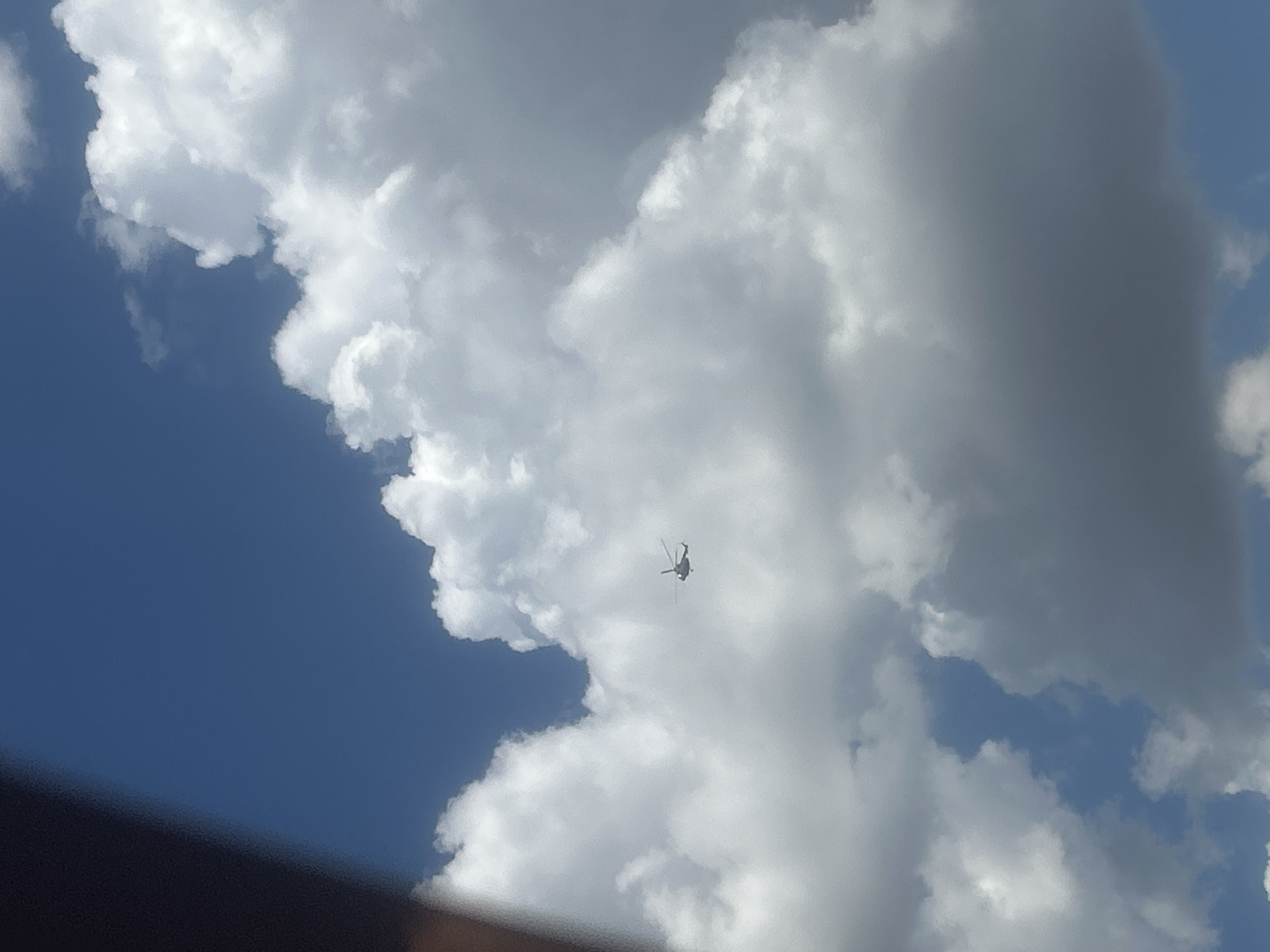 A single helicopter is visible in the sky amidst white and gray clouds. The helicopter appears to be a light-colored model. The sky is blue with scattered clouds of varying densities. A dark, out-of-focus object is present in the lower left portion of the frame, partially obscuring the sky. The scene is an outdoor environment.