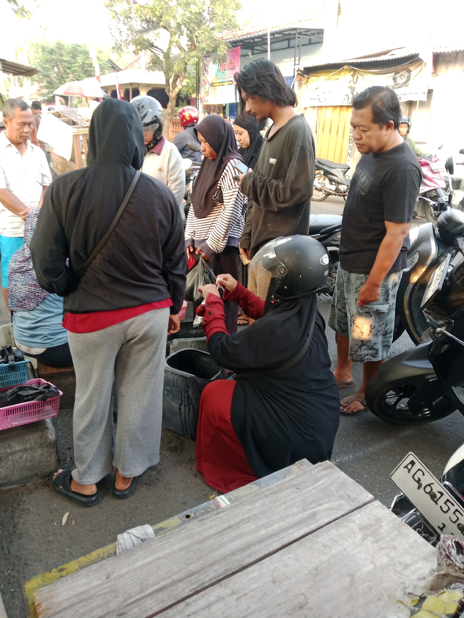 The image depicts an outdoor scene in Kediri City, Indonesia, likely a street or market area, with several individuals present. In the foreground, a person wearing a black hooded jacket and grey sweatpants is facing away from the camera, with a black crossbody bag. To their left, a person in a blue and white patterned shirt is partially visible, seated. A woman in a black hijab and red top is kneeling and appears to be engaged in an activity with something held in her hands. She is wearing a black helmet. To her right, a man in a black t-shirt and camouflage shorts stands looking down, holding a small, light-colored object. Behind these individuals, other people are visible, including a man in a white shirt and a woman in a striped shirt and black hijab. Motorcycles are present in the background, suggesting a public thoroughfare. The environment appears to be a slightly dusty, paved area with some vegetation and buildings in the distance. A wooden structure with a yellow stripe is in the lower part of the frame. A license plate with the characters "AG.6155" is visible on a motorcycle in the right foreground.