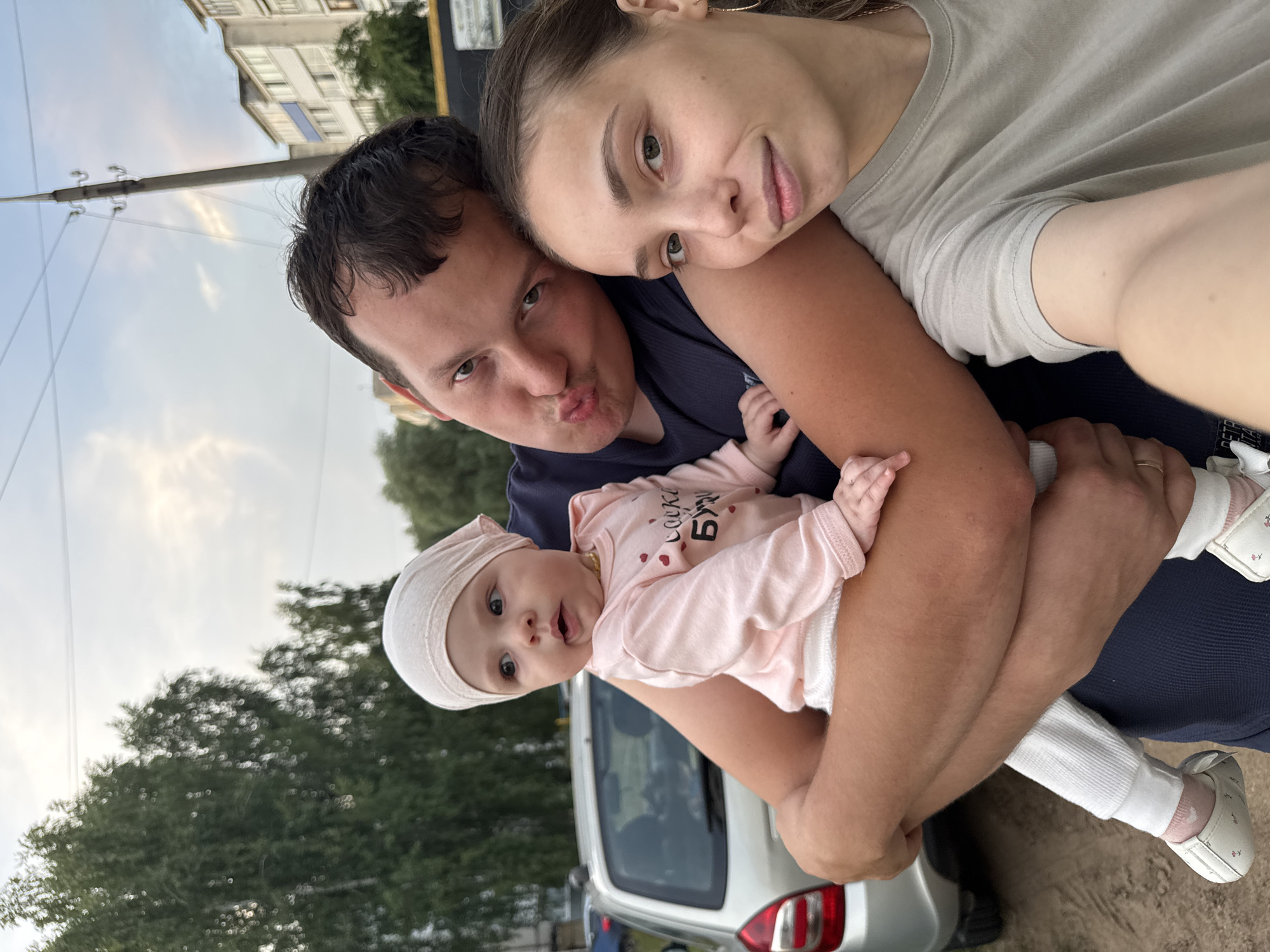 A family of three, a man, a woman, and a baby, pose for a selfie outdoors. The man is in the foreground, holding the baby in his arms and making a duck face. The woman is to his right, resting her head on his shoulder. The baby, wearing a pink onesie and a matching headscarf, looks to the side with a surprised expression. The background shows a street with a car, trees, utility poles, and buildings, suggesting an urban or suburban setting in Noviy, Russia. The lighting and cloudy sky indicate it is likely late afternoon or early evening.