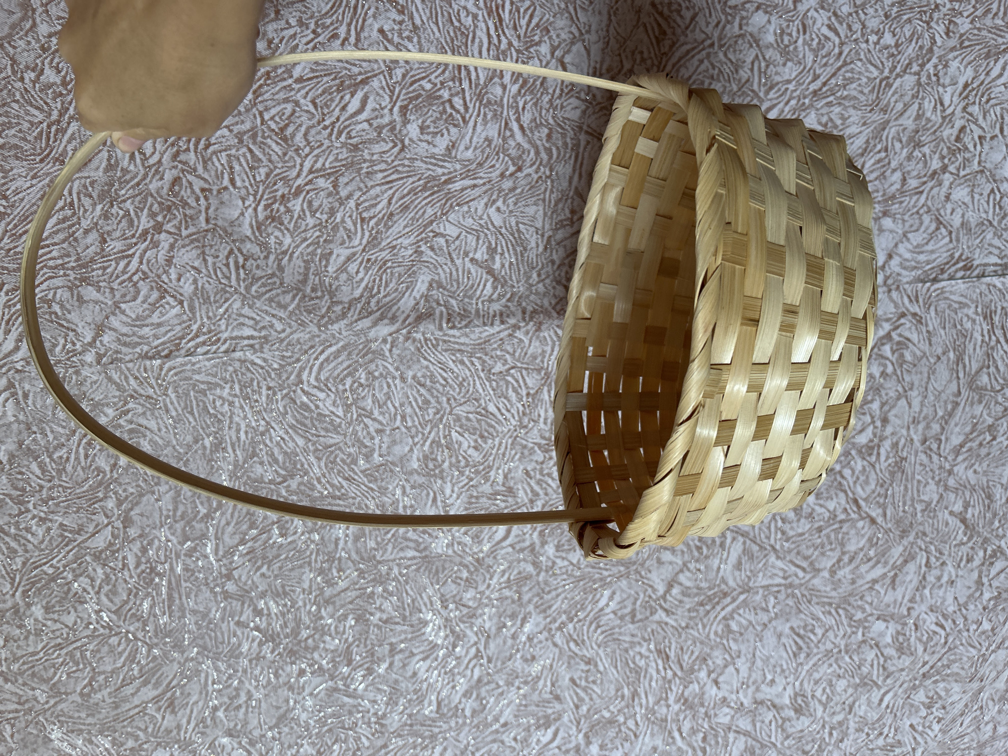 A close-up, eye-level shot shows a light brown woven bamboo basket with a curved handle being held by a person's hand. The basket is empty and hangs against a textured, light-colored wallpaper with a crinkled pattern and scattered glitter-like highlights. The hand holding the basket is positioned in the upper right corner of the frame, with the fingers subtly gripping the handle. The background wallpaper exhibits a consistent pattern across the image, with no discernible environmental features to indicate a specific city or location like Gelendzhik, Russia.