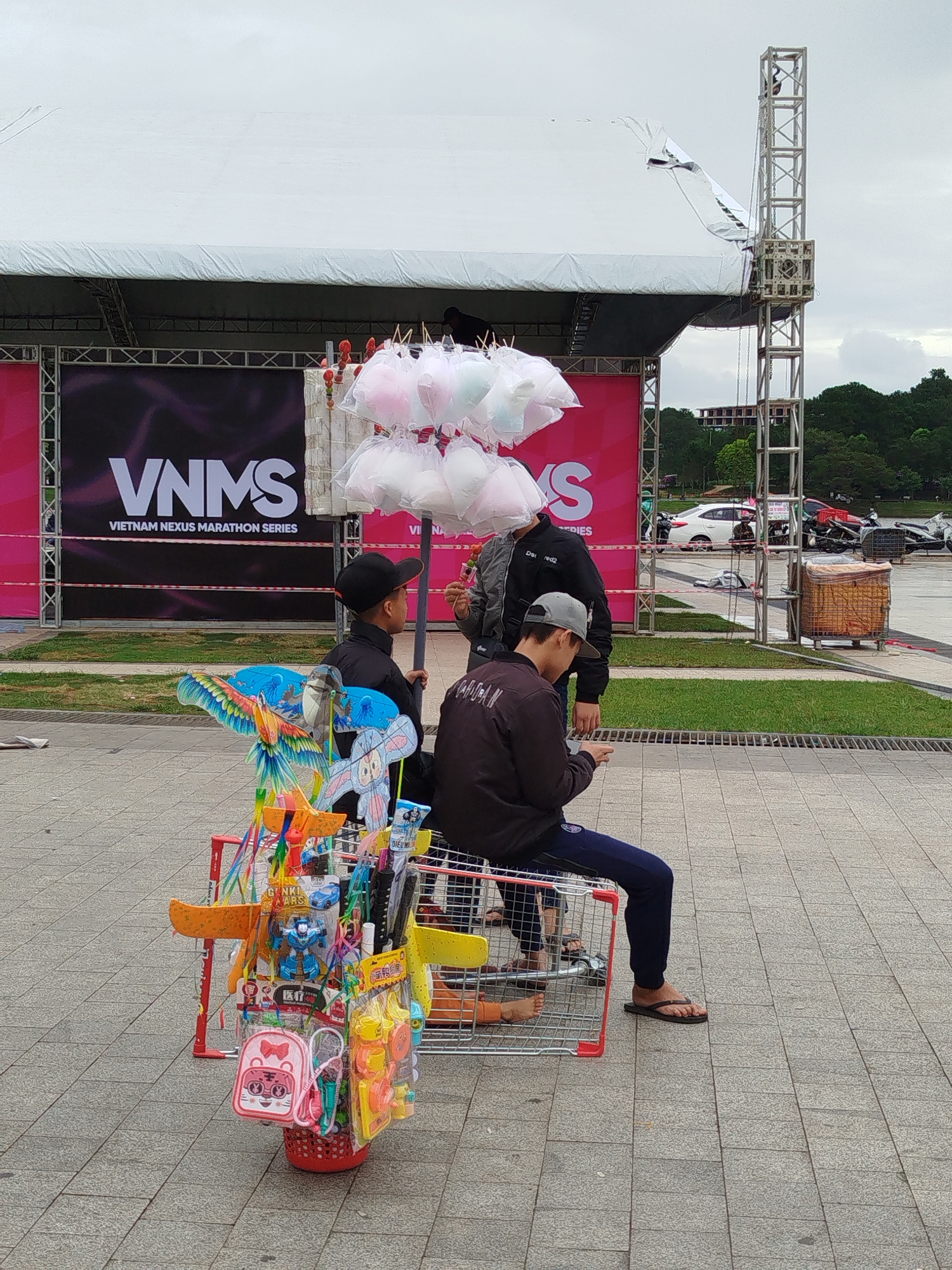 A street scene, likely in Da Lat, Vietnam, shows three males gathered near a cart filled with toys and cotton candy. A VNMS (Vietnam Nexus Marathon Series) banner forms the backdrop. A young boy in a black cap and jacket is standing by the cart. An adult wearing a grey cap and brown jacket sits on the cart, looking at a mobile device in his hand. A third male in a black jacket is standing in the middle. The cart holds colorful kites, toys, and a red basket full of items. Above them, bags of cotton candy are displayed. Behind them, there is some parked cars and motorcycles. A metal structure rises in the background. The scene takes place on a paved area.