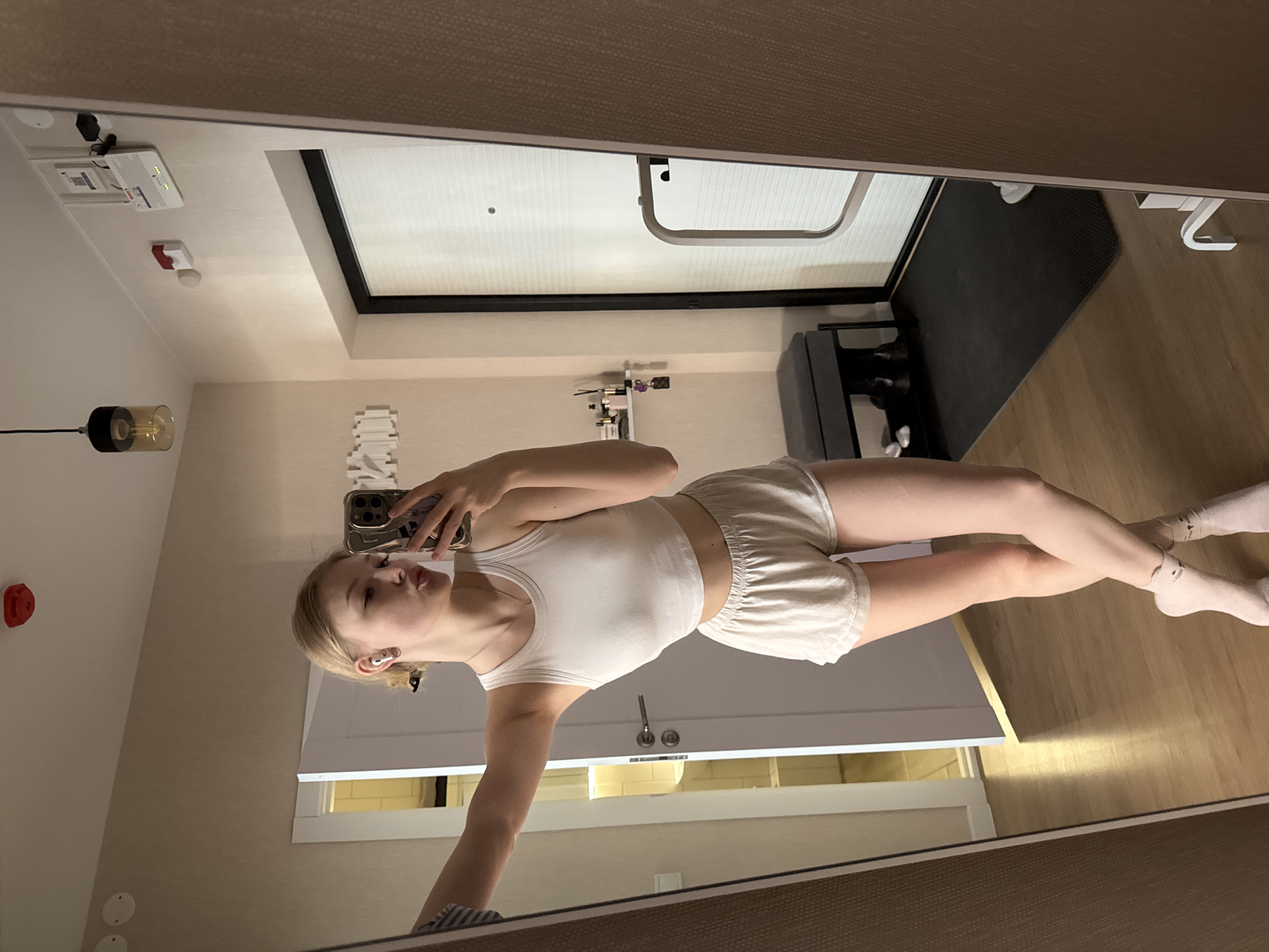 A young woman with blonde hair is taking a selfie in a mirror. She is wearing a white tank top and matching shorts. She has a phone in her left hand. She is wearing white socks. The room appears to be an apartment with a door leading to what might be a bathroom or closet. The reflection in the mirror includes elements of a modern interior, such as a light fixture, and a small black workout mat. There are no elements to confirm the location. It's a personal image of a woman taking a selfie in an apartment.
