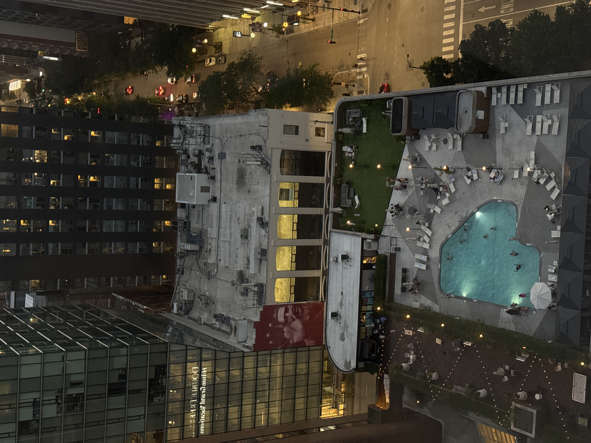 This image depicts an aerial view of a rooftop pool and lounge area at the Hotel Julian in Chicago, USA. The scene includes a turquoise swimming pool with several individuals swimming and relaxing, surrounded by lounge chairs and umbrellas. Adjacent to the pool is a grassy area with more seating and umbrellas.
In the background, a large building with a mural of a man's face is visible. This is part of the Hotel Julian, located at 168 N. Michigan Avenue. The surrounding urban environment of Chicago is characterized by tall buildings, a street with traffic and streetlights, and trees lining the sidewalks. The time of day appears to be dusk or early evening, as indicated by the artificial lighting from the buildings and street lamps.