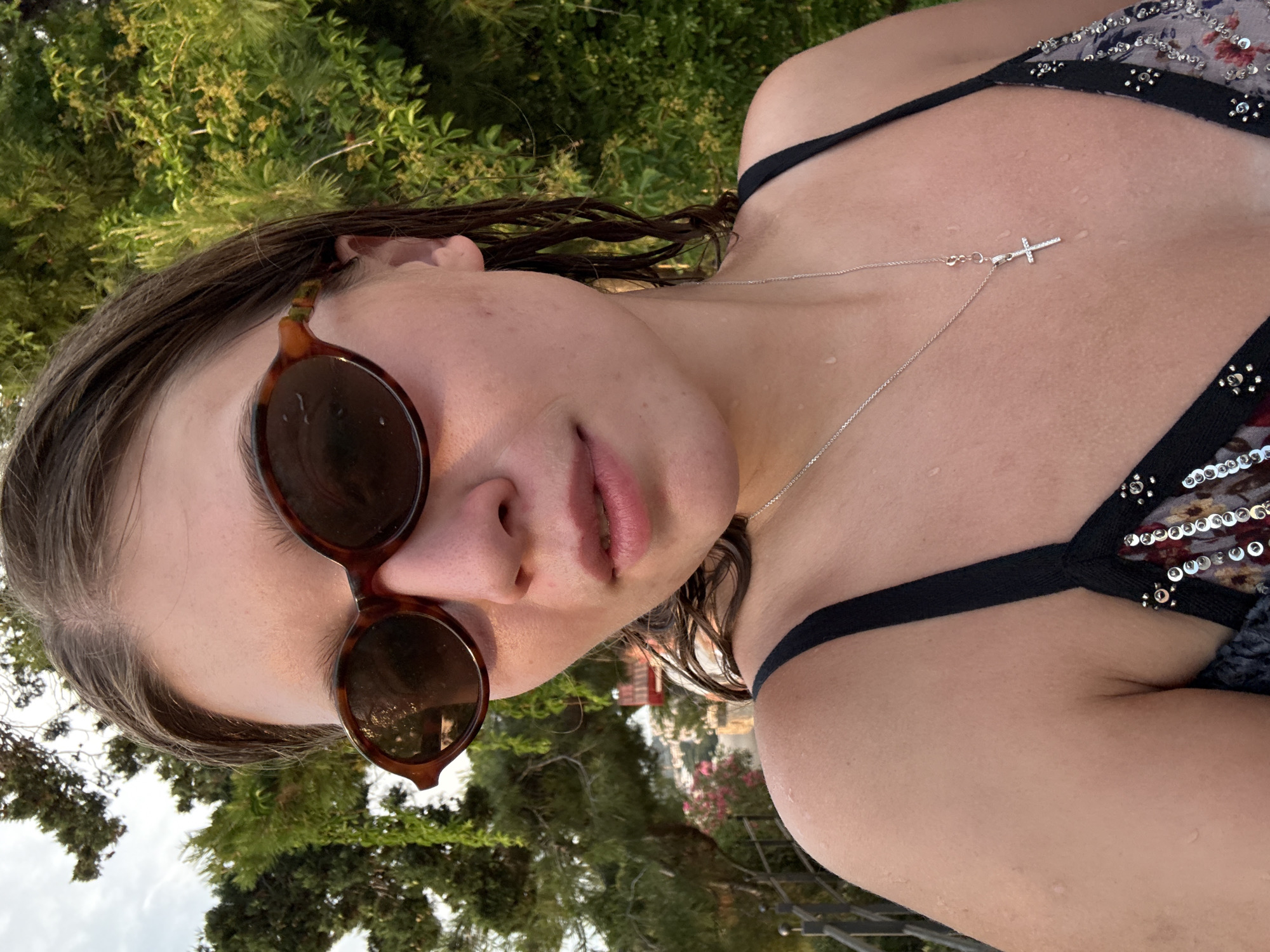 A young woman wearing sunglasses and a bikini poses for a selfie in Primošten, Croatia. The woman has wet brown hair and is wearing a silver necklace with a cross pendant. She is standing outdoors with lush green foliage and trees in the background. The sky is visible above the trees.