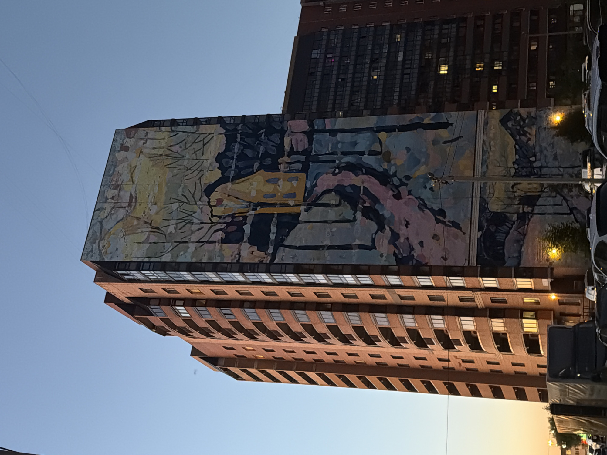 A tall, brick apartment building with a large mural on its side dominates the frame, set against a clear, pale blue sky. The mural depicts a stylized landscape with a yellow house, bare trees, and a winding path. To the right of the muraled building is another, darker apartment building with many lit windows. In the foreground, the tops of several cars and a covered truck are visible, suggesting a street scene. The light in the sky indicates it is either early morning or late evening.