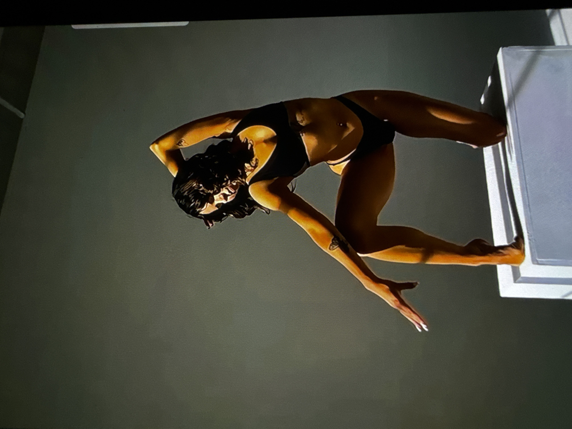 An individual, appearing to be a female with dark, wavy hair, is depicted posing in an indoor studio-like environment. She is wearing a dark, two-piece bikini or athletic underwear set, and has a visible belly button piercing. Tattoos are present on her left upper arm (appearing as a geometric or abstract symbol), her right forearm (resembling a butterfly), and a small marking on her left torso. Her right hand, extended forward with fingers spread and featuring white-tipped nails, rests near a white, rectangular platform. She is kneeling on her left knee on this platform, with her right foot also on the platform, balanced on her toes. Her left arm is bent, raised overhead. The scene is illuminated by strong, directional lighting from the upper left, casting sharp shadows and highlighting the subject's skin with a warm tone against a plain grey background. The image appears to be a photograph of a digital screen due to visible pixelation and black borders on the right and top edges. There are no discernible visual elements within the image to identify the location as the city of Varna, Bulgaria, or any specific geographical location.