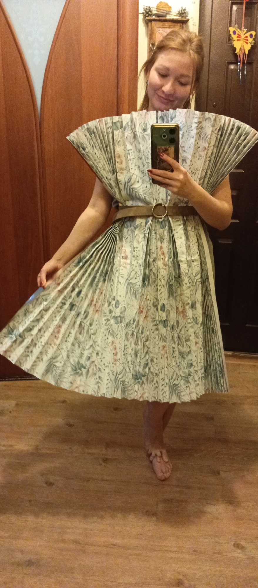 A full-length indoor shot shows a fair-skinned woman posing while holding a smartphone in her right hand to take a selfie in front of a wooden door. The woman is wearing a paper dress with pleated sections and a light brown belt with a circular buckle cinching the waist. The dress features a floral pattern with shades of green, tan, and light blue. She is barefoot and looks towards the camera with a slight smile. Her hair is pulled back, and she has a light complexion.
The background includes a wooden floor and a wooden door with curved panels on the left. Behind the woman, a darker wooden door has decorative elements, including a hanging butterfly ornament, and a partial view of a clock is visible above the door. The lighting is soft, and the overall atmosphere is casual.