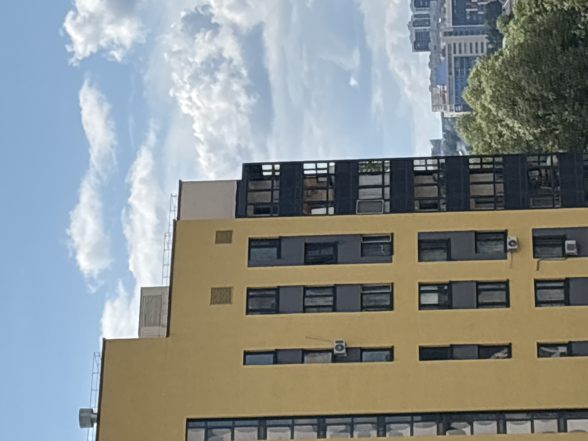 An exterior, eye-level, vertical shot shows a close-up of a modern, yellow apartment building with dark-framed windows. The building dominates the left side of the frame, with a few air conditioning units visible. To the right of the apartment building, other buildings of varying heights and colors are visible in the distance, along with lush green trees. The sky is a vibrant blue, dotted with fluffy white clouds. The scene appears to be a typical urban landscape, likely in the city of Kazan, Russia.