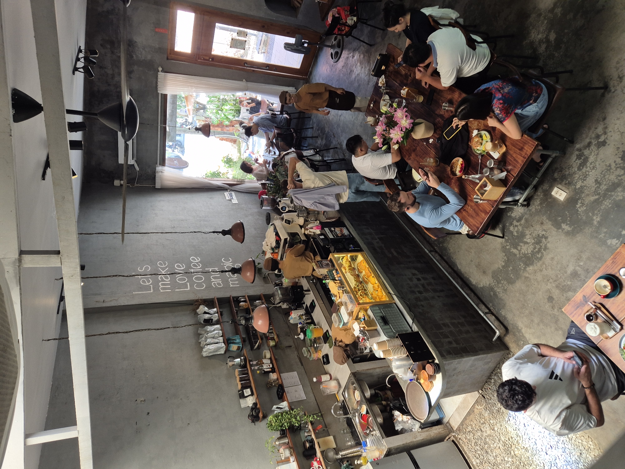 This image shows the interior of a modern, industrial-style cafe with a concrete wall and exposed ceiling beams. The cafe is populated with several patrons seated at tables and standing at the counter. A barista is seen working behind the counter, preparing drinks. The cafe is decorated with plants and a mural on the wall that reads "Let's make Love coffee and magic." The overall atmosphere is casual and inviting, suggesting a popular spot for locals and tourists alike. It is likely located in Đà Nẵng, Vietnam, a coastal city known for its vibrant cafe culture and modern aesthetic.