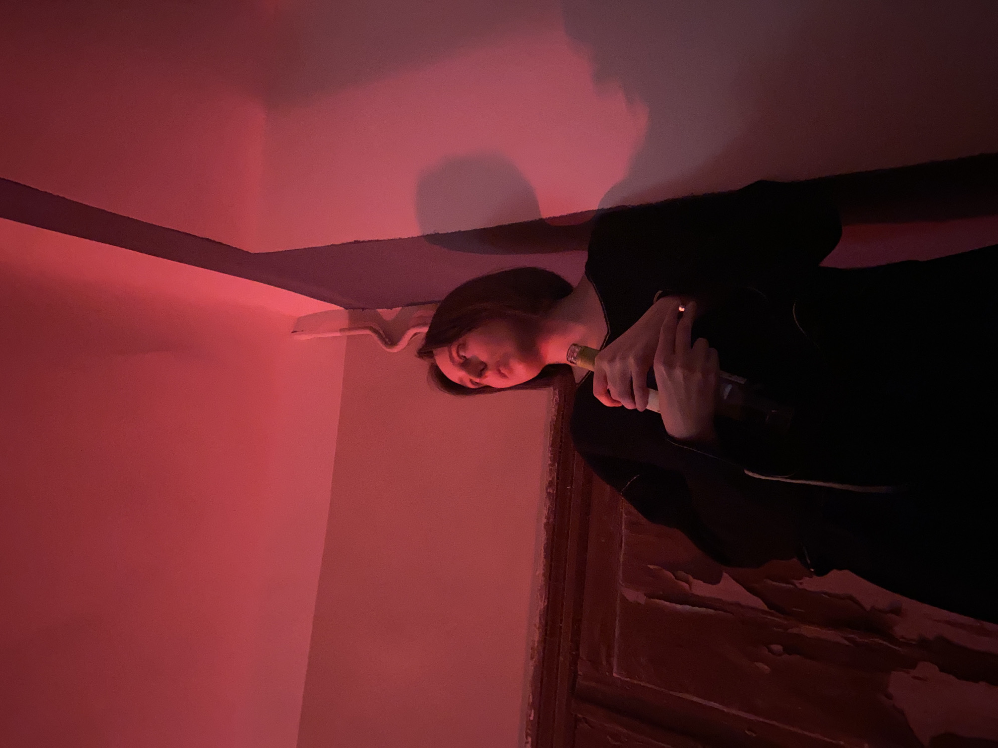 A woman is holding a bottle and a lit cigarette inside a room. The walls are painted red. A door with a rough surface is located near her. The lighting is dim. While a location is mentioned in the prompt (Moscow, Russia), no specific details in the image corroborate or negate this claim.