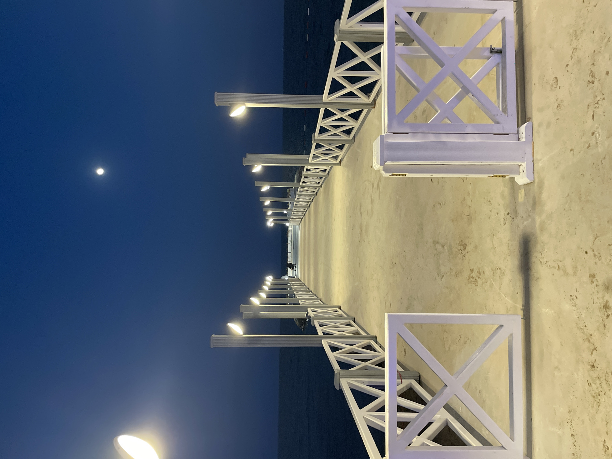 The image depicts a long, illuminated pier extending over a dark body of water, likely the Mediterranean Sea, at dusk or night. The pier is lined with white lampposts that cast a warm glow on the concrete walkway. A white, decorative railing with crisscross patterns runs along both sides of the pier. The sky is a deep twilight blue with a small, bright moon visible. In the distance, at the end of the pier, two figures are present, one appearing to be seated. The pier is located in the vicinity of Trikomo, Northern Cyprus. The scene conveys a sense of calm and serenity.