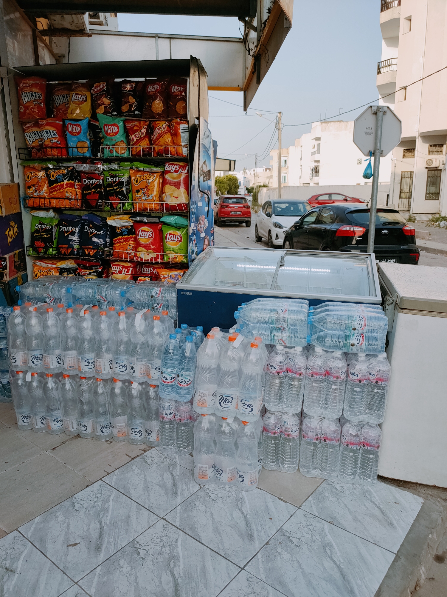 This media file shows an outdoor display of snacks and bottled water in front of a store. The primary subjects are shelves stocked with various brands of chips, including Lay's, Doritos, Bugles, and Takis. Below the snack shelves, numerous cases of bottled water are neatly stacked. To the right of the snacks, there's a large, open-top freezer, also filled with bottled water, and a white refrigerator unit.
The setting appears to be a street or sidewalk in front of a small business, possibly a convenience store or market. In the background, there are buildings, a street with several cars parked and driving, and power lines. The cars visible are a red hatchback, a white hatchback, and a black Kia hatchback. A prominent white octagonal sign with "MJS" printed on it is visible on a pole.
The time of day appears to be late afternoon or early evening, indicated by the soft, warm light and the long shadows. The weather seems clear and pleasant. There are no people visible in the image.
Visible text includes brand names on the snack packaging like "Bugles," "Lay's," "Doritos," and "Takis." The bottled water labels show brands like "Marwa," "Safia," and "Mira." Text on the white refrigerator unit reads "UGUR." The octagonal sign has "MJS" and a license plate is visible on the black car with the text "88 3187".
The overall impression is that of a commercial establishment set up to sell beverages and snacks to passersby, likely in a moderately busy urban or semi-urban area.