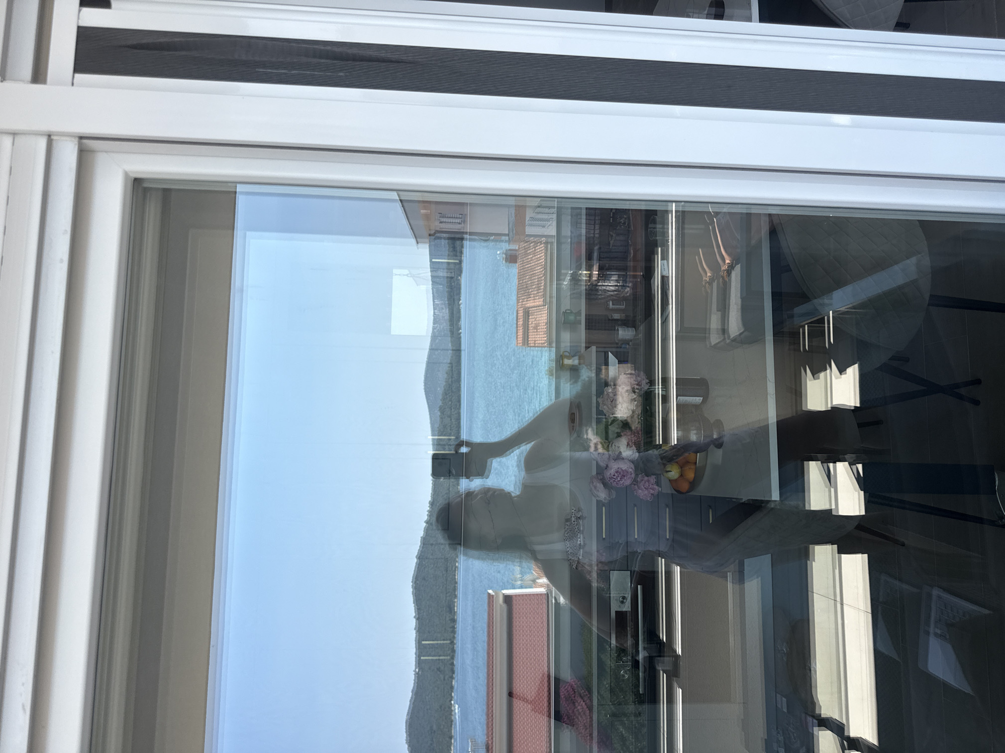 This is a photograph taken from a window, likely in Šibenik, Croatia, as suggested by the location context. The image is oriented vertically. It shows a reflection on the window, with the actual scene visible through the glass.
In the reflection, a person is seen taking a selfie with a smartphone. They are positioned in what appears to be an interior space, with a table in front of them holding a vase of flowers and a bowl of fruit. A dining table and chairs are also visible in the reflection. The interior space seems to be a well-lit room, possibly a dining area or living room, with windows and furniture.
Through the window, a scenic view of the Adriatic Sea and a coastline dotted with hills and buildings is visible. The water is a vibrant blue, and the sky is clear and bright, suggesting a sunny day. The architecture in the background, with terracotta-tiled roofs, hints at the Mediterranean style typical of the region.
The overall impression is one of a bright, pleasant day in a coastal location. The activity of taking a selfie suggests a personal moment of capturing the experience. The scene combines an indoor setting with a beautiful outdoor landscape, viewed through the reflective surface of a window.