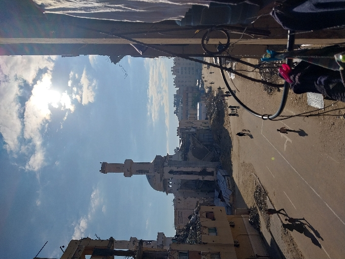 A damaged mosque with a tall minaret stands amidst the rubble of destroyed buildings under a bright sky. People walk along a street marred by destruction. The scene is observed from a high vantage point, possibly a damaged building. Laundry hangs on a balcony railing in the foreground. The surrounding area appears devastated, bearing witness to conflict or disaster.