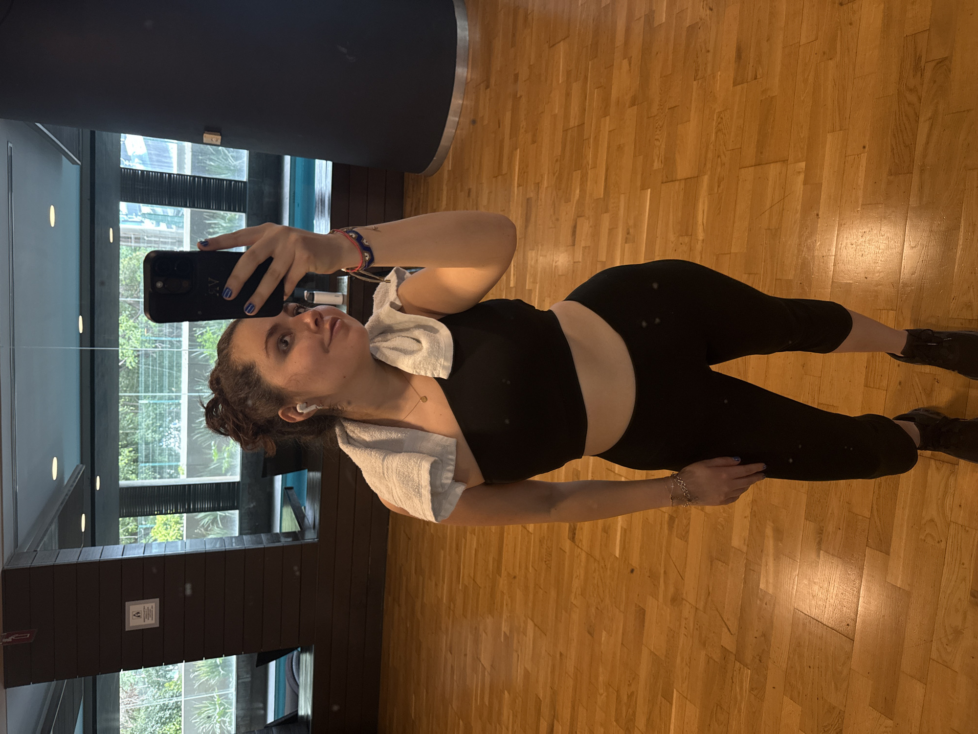 A woman with curly dark hair, wearing a black workout outfit, including a tank top and leggings, is taking a selfie in a gym. She holds a black smartphone with her left hand, showing blue nail polish. A white towel is draped over her left shoulder. She's wearing a light-colored necklace and a bracelet on her right wrist. The gym has a wooden floor and a large window looking out onto greenery and buildings. The window frame appears to be made of dark wood. A sign is visible on the wall beside the window. The location appears to be Mexico City, Mexico, based on the architecture and vegetation visible through the window.