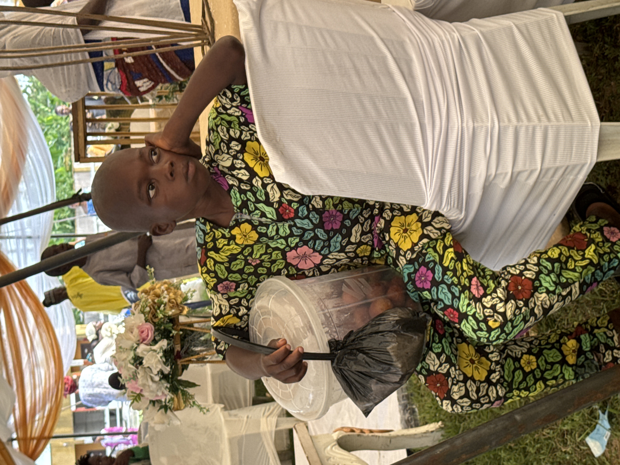 A young boy with a shaved head sits on a white chair with draped fabric, holding a clear plastic container with a black lid filled with round, reddish-brown objects, likely fruit. The container has a black handle to which a black plastic bag is tied. The boy wears a colorful, floral-patterned outfit in shades of black, green, yellow, pink, and red. His left elbow rests on his hand, and his gaze is directed away from the camera. In the background, a floral arrangement sits on a wooden stand. Other people, some wearing traditional attire, are visible, and the scene appears to be set up under a tent with draped orange and white fabric. The environment suggests an outdoor gathering or event. Based on the context of the event and typical scenery in Badagry, Nigeria, the setting is likely an outdoor gathering within the city, possibly a market or a community event.