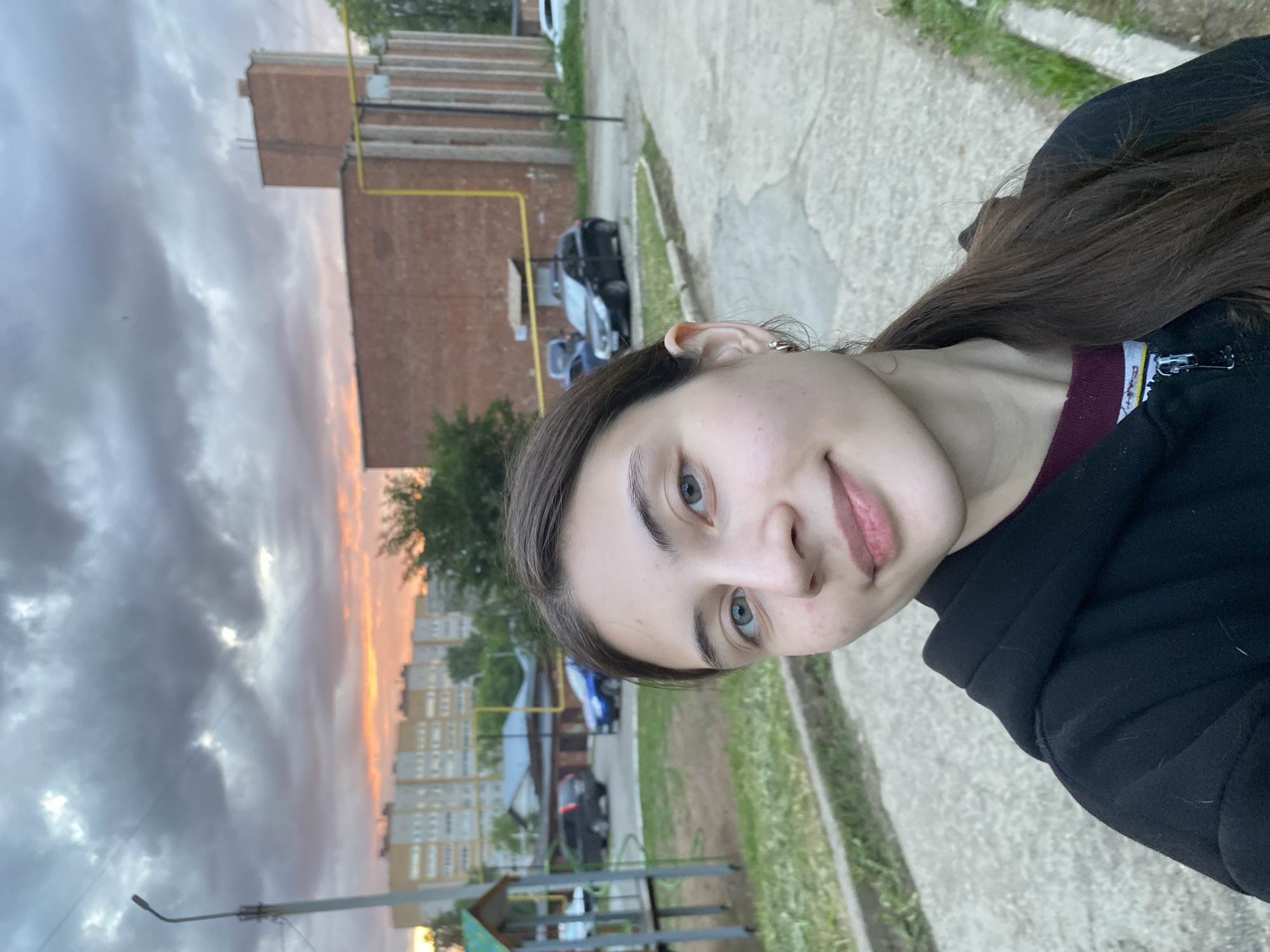 The image shows a young woman taking a selfie. She is standing on a paved surface with grass to her right. Behind her are buildings of different types: a multi-story apartment building, a brick building, and other structures. Cars are visible in the background. The sky is cloudy with a hint of sunset. The location appears to be Votkinsk, Russia, based on the urban environment and the presence of buildings typical of Russian cities. A playground or park structure is partially visible in the foreground.