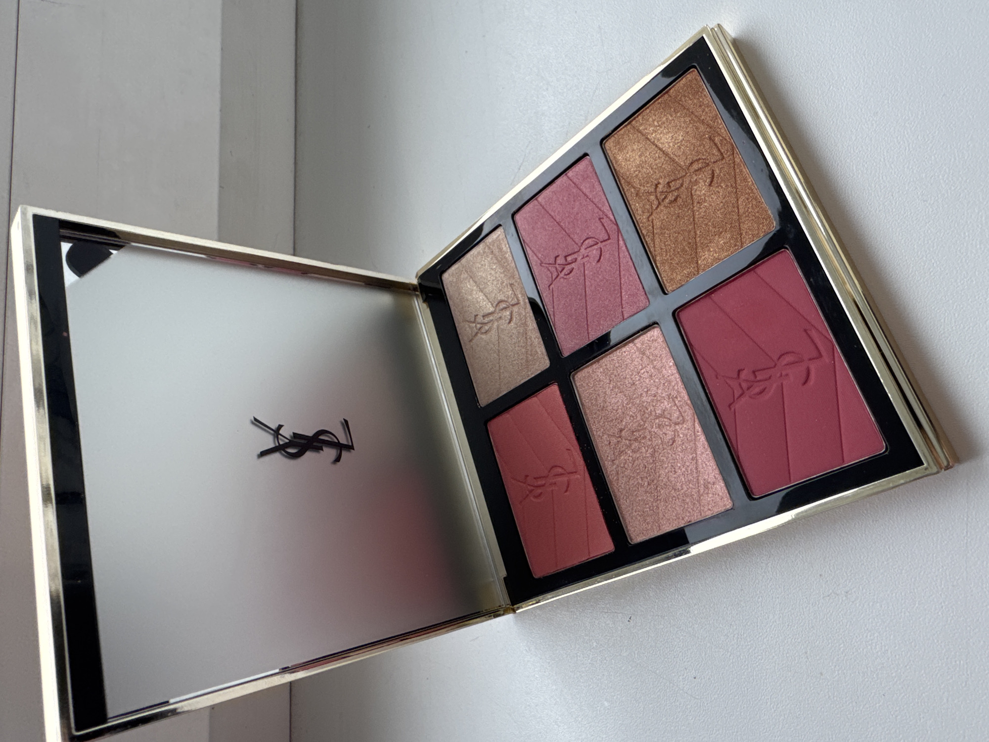 A close-up shot depicts an open YSL (Yves Saint Laurent) beauty palette containing six different makeup shades. The palette is gold-colored with a black interior housing the makeup compartments. The shades vary in color, from light gold, rose gold, and pink to deeper burgundy and bronze tones. Each pressed powder shade features the YSL logo imprinted into it. The palette's lid, opened to the side, has a mirror-like finish with a black YSL logo in the lower center. The surface below the palette is a white or light gray color, suggesting a table or counter. The backdrop consists of an off-white or light gray wall. The location is likely indoors, possibly in Ждановіцкі сельскі Савет, Belarus.