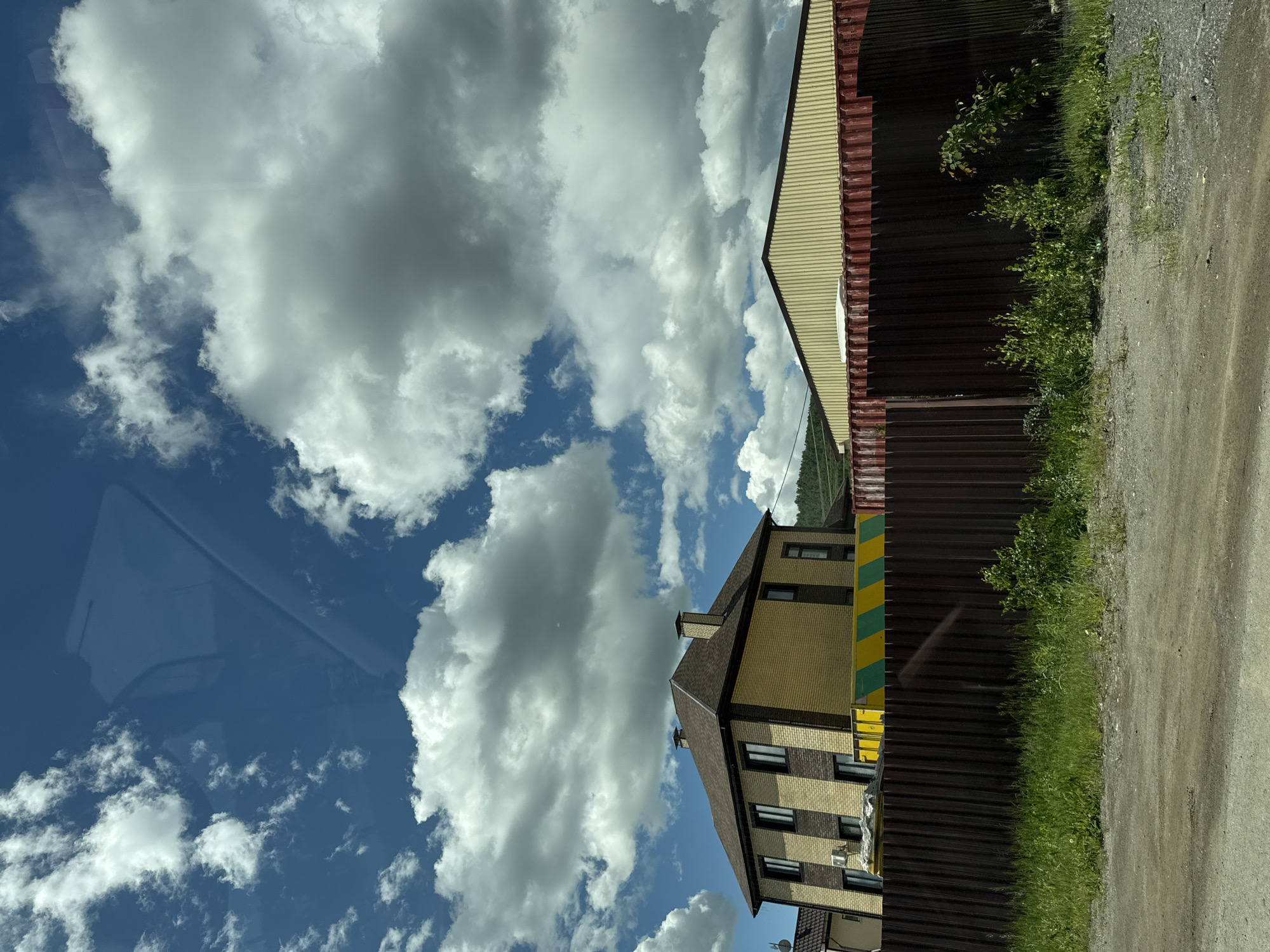 The image depicts a residential and industrial scene in Uchaly, Russia, under a sky filled with dramatic cumulus clouds. In the foreground, a dirt road transitions into a strip of green grass bordering a dark brown corrugated metal fence. Behind the fence, a two-story brick house with a brown shingled roof and dark-trimmed windows stands to the left. The house is primarily yellow brick with darker vertical brick accents. To the right of the house, a large, long, yellow building with a ridged roof is visible, partially obscured by a section of red shipping containers. The overall impression is a semi-rural or developing area, possibly on the outskirts of the city.