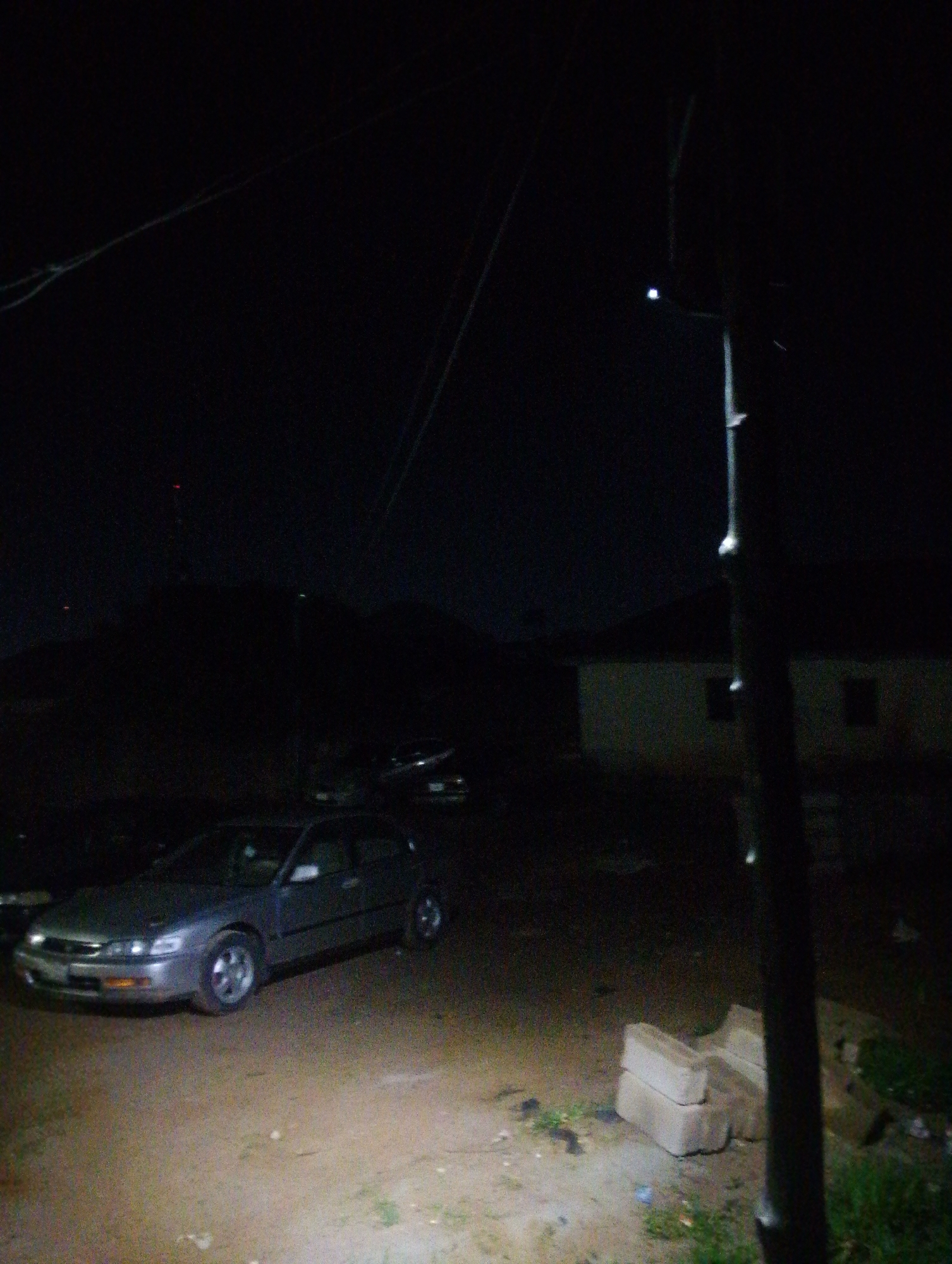 The image captures a dark, unlit outdoor scene in Jos, Nigeria, likely at night. A grey sedan is parked on a dirt or gravel surface, illuminated by an unseen light source from the right, casting shadows. The car appears to be a Honda Accord. In the background, the silhouette of a house with a dark roof and windows is visible. A tall, dark pole, possibly a streetlight or utility pole, stands prominently in the right foreground, with some faint lights or reflections on it. Overhead, power lines are faintly visible against the dark sky. A small stack of what appear to be bricks or concrete blocks lies on the ground to the right of the car. The overall impression is one of a quiet, possibly residential area at night.