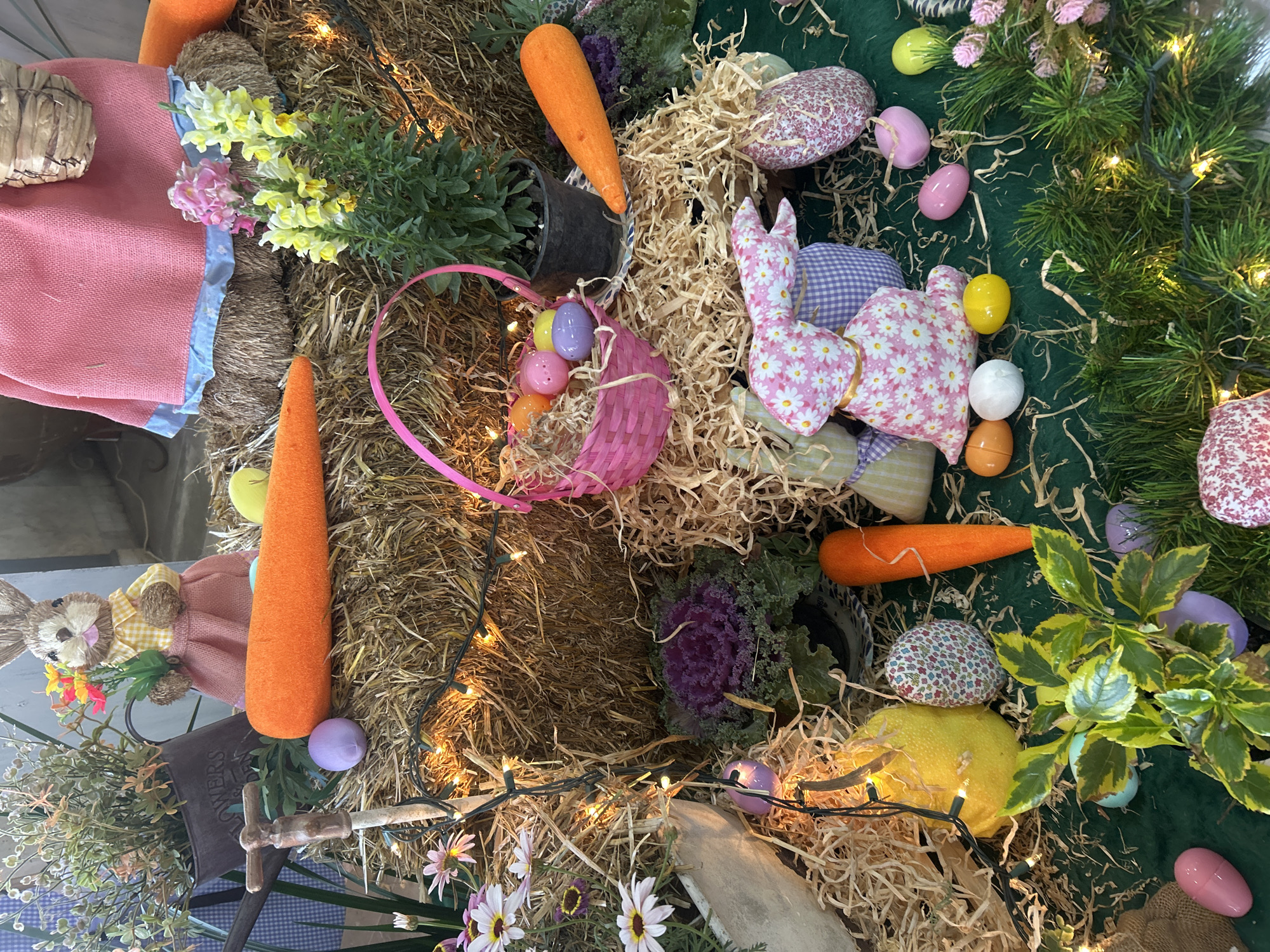 The image captures a whimsical, overhead view of an Easter-themed display. The scene is composed of straw bales adorned with various Easter decorations. There are stuffed bunnies crafted from floral-patterned fabric, interspersed with faux carrots and colorful Easter eggs. A small pink basket contains additional eggs. Light strings are interwoven throughout the straw, providing soft illumination. The composition also includes live plants, some in pots. The environment suggests a staged or decorated space, possibly for a seasonal event or display, and is confirmed to be taken in Puebla, Mexico.