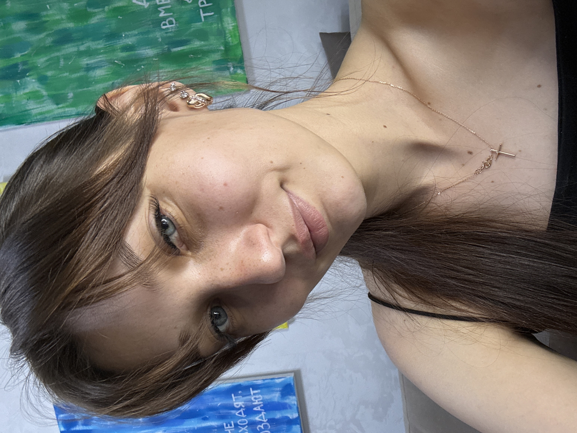 A young woman with brown hair and blue eyes is shown in a close-up portrait. She is wearing a black camisole and a rose gold necklace with a cross pendant. The background features abstract paintings, one blue and one green. The woman is indoors, possibly in a home or studio environment. There are no recognizable landmarks or any elements that would allow for identification of the location in Novokuznetsk, Russia. The scene is well-lit and the woman is looking directly at the camera.