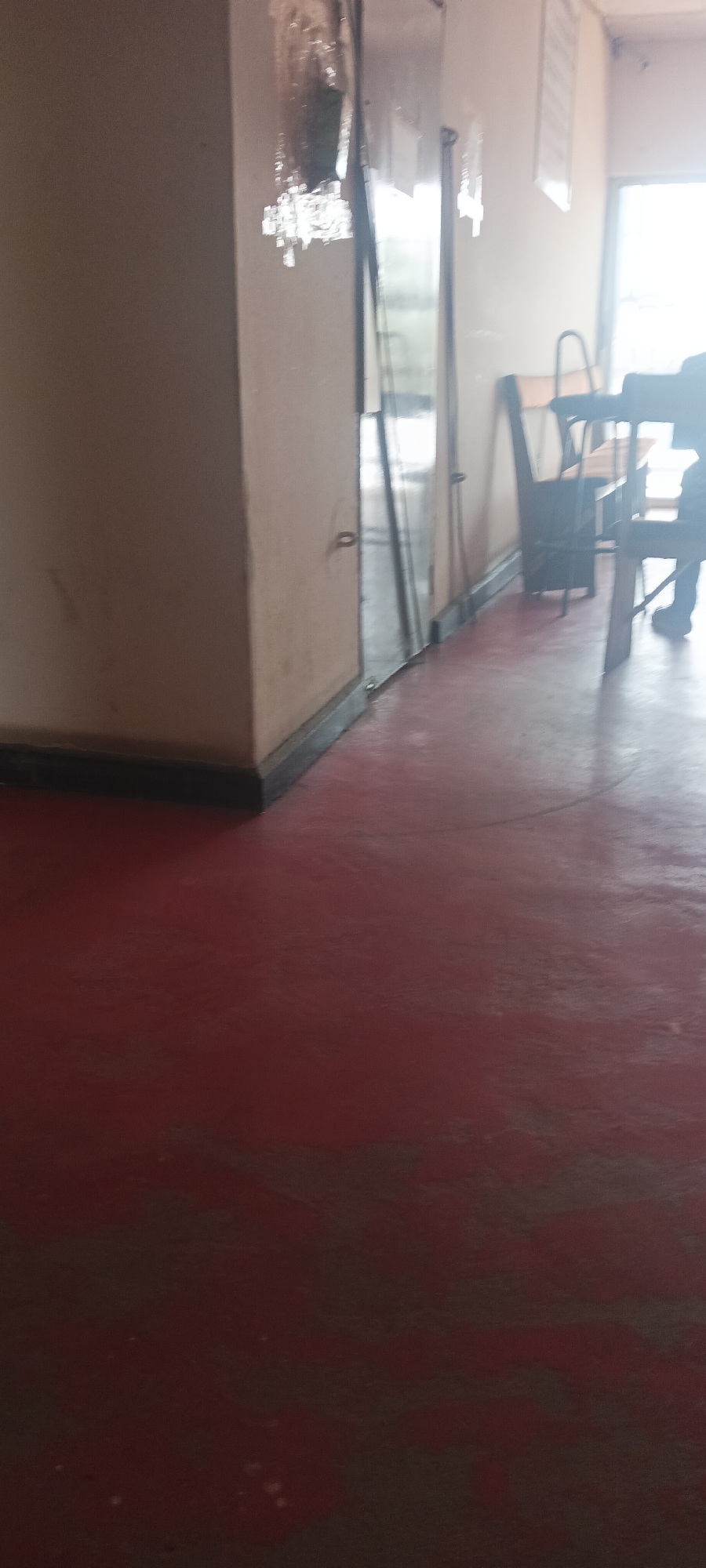 The image depicts an interior space, possibly a dining area, in what may be a commercial establishment in Jos, Nigeria. A red-painted concrete floor dominates the foreground. Adjacent to the left, a beige-painted wall extends vertically, contrasted by a black baseboard. To the right of the wall is a glass-paned partition with a metal frame. In the background, a blurred view of tables and chairs suggests a dining setup. A person, partially visible, is seated at one of the tables. The overall lighting appears bright and diffused, indicating daylight conditions. There are no immediate signs of forensic evidence.