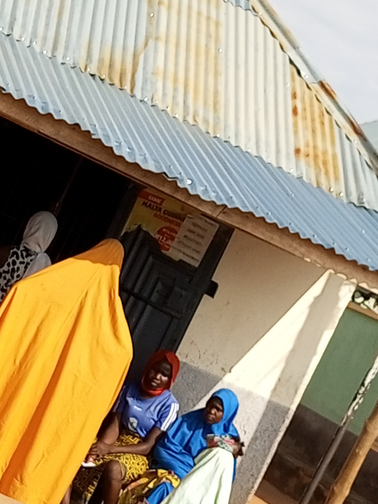 An outdoor scene in Gombe, Nigeria, shows a corrugated metal roof, part of a building's structure. Below the roof, two women are seated. One woman, on the right, wears a blue headscarf and a blue t-shirt with white stripes on the sleeves. She holds a baby or a bundle wrapped in a light-colored cloth. The second woman, to her left, has a red headscarf and a blue t-shirt with white stripes. She is seated next to the first woman. To the left of these seated women, a third person is visible, draped in an orange cloth, obscuring their features. Further to the left, another individual is partially visible, wearing a white head covering and a black and white patterned garment. Behind the seated women, a dark doorway is visible, with some signage on the wall above it. The ground appears to be a mixture of dirt and possibly some pavement. The overall lighting suggests it is daytime.