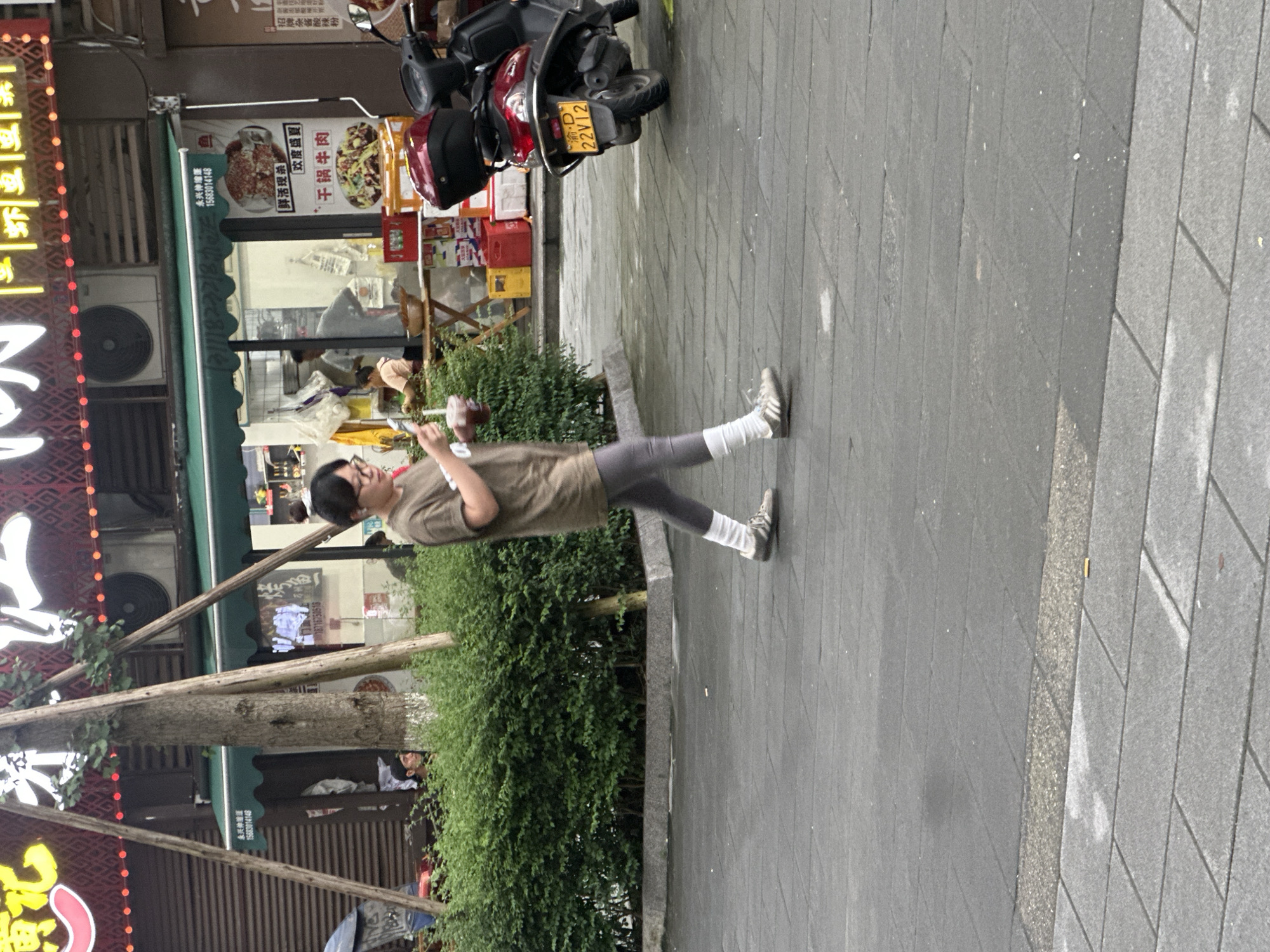 A person wearing glasses, a brown t-shirt, gray leggings, and white socks is walking on a sidewalk. They are holding a light-colored object in their right hand. The person is walking away from the camera. In the background, there is a building with awnings and signs, a motorcycle, and a bush. One sign in the background appears to be for a restaurant with images of food. The ground is paved with gray tiles. The overall scene appears to be a street in a city, possibly in an urban commercial area. The lighting suggests it is daytime.