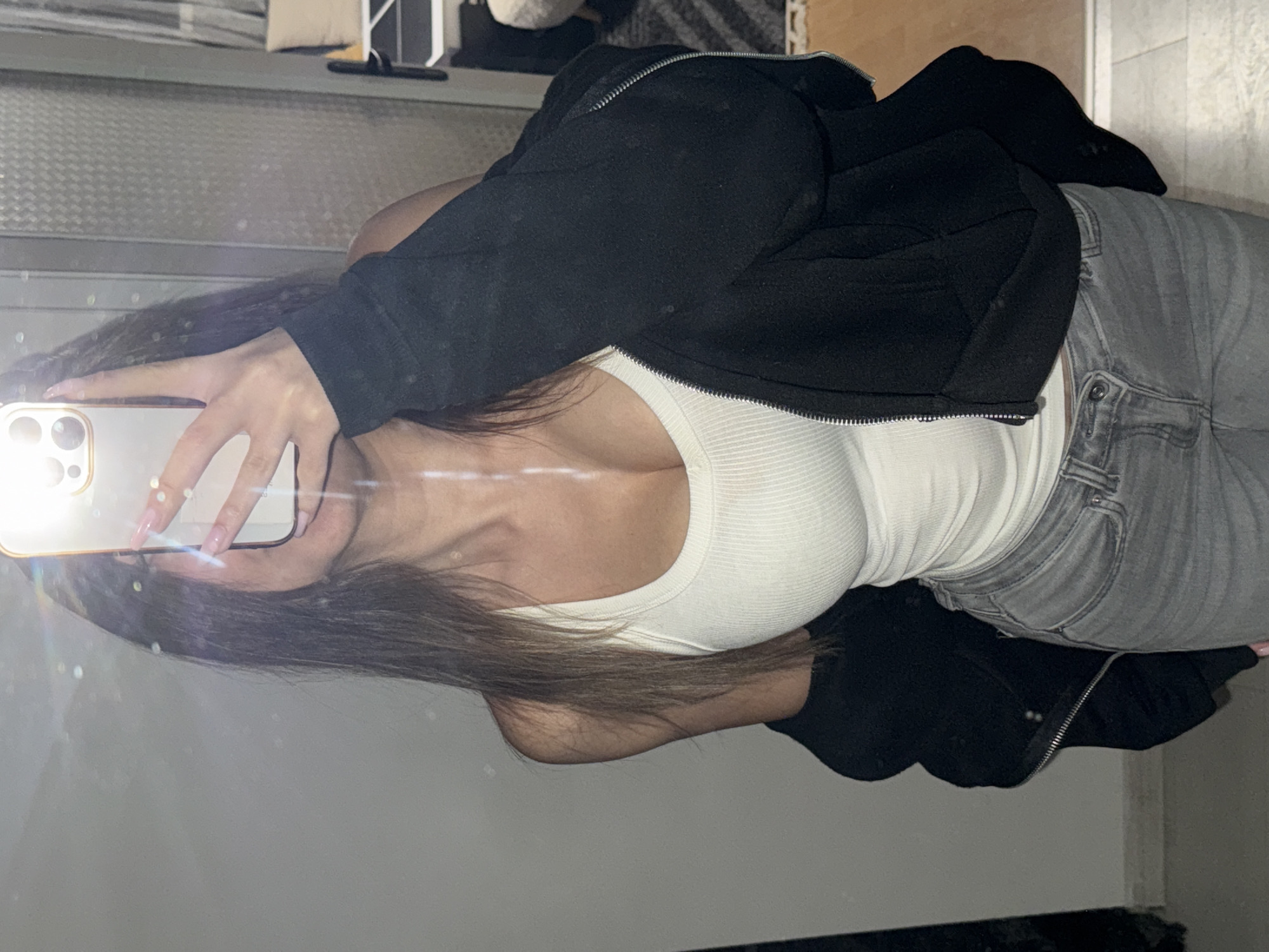 An indoor mirror selfie captures a person from the chest up, with their face obscured by a smartphone. The image is taken at a slight angle and appears rotated clockwise, showing the subject sideways. The person holds a light-colored smartphone with multiple rear cameras, from which a bright flash emanates, creating a prominent lens flare effect. Long, dark brown hair flows over the shoulders and chest.
The individual is dressed in casual attire, featuring a black zippered hoodie or jacket worn open over a fitted, white ribbed tank top or camisole. They are also wearing light grey jeans. The reflection in the mirror reveals an indistinct indoor setting, possibly a room with muted colors and subtle patterns in the background, suggesting a home or private space. No specific time of day or emotions are discernible, and there is no visible text. The location context provided is Sofia, Bulgaria.