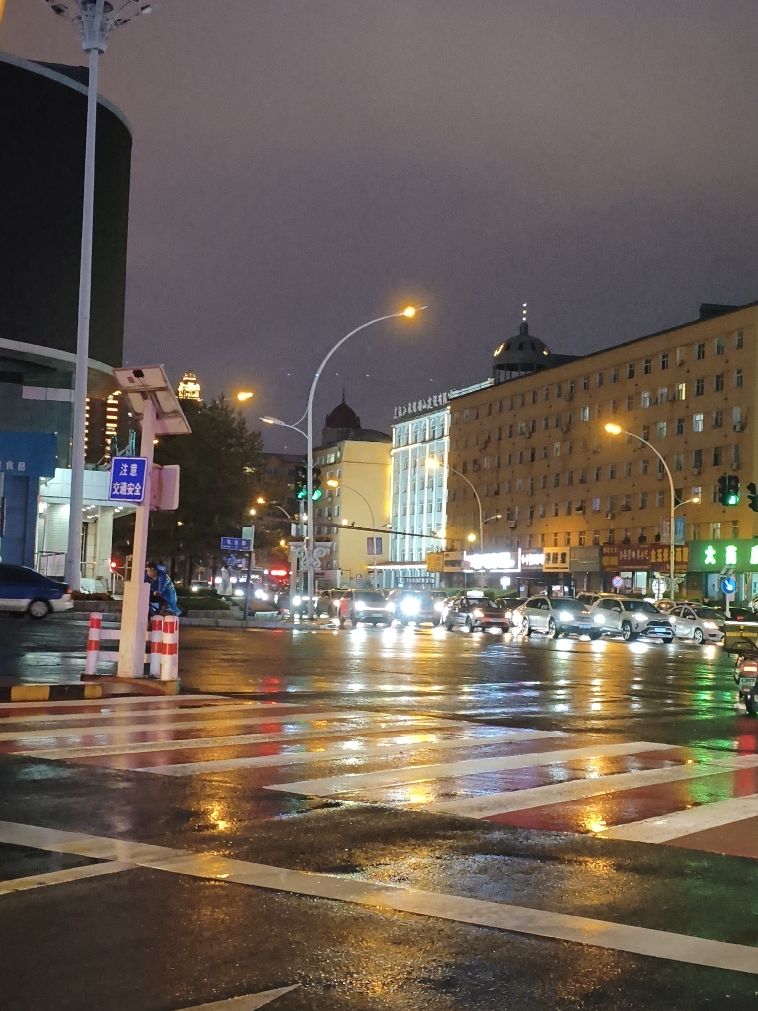 This is a photograph taken at night in Jixi, China. The scene depicts a wet street with traffic lights and vehicles. The road surface is reflective, showing the lights of cars and streetlights. In the foreground, a pedestrian crossing with white and red stripes is visible, glistening with moisture. Several cars are stopped at the intersection, their headlights illuminated. Traffic lights are showing green for vehicles heading forward.
To the left of the image, there is a tall lamp post and a sign that reads "注意 交通安全" (Zhùyì jiāotōng ānquán), which translates to "Attention: Traffic Safety." A person in a blue jacket is standing near this sign, leaning against a pole.
In the background, there are multi-story buildings. One building on the right is a yellowish-brown color with many windows. Another building on the left is lighter in color. The sky is overcast and dark, contributing to the nighttime atmosphere. The overall impression is a city intersection at night during or after rainfall, with active traffic.