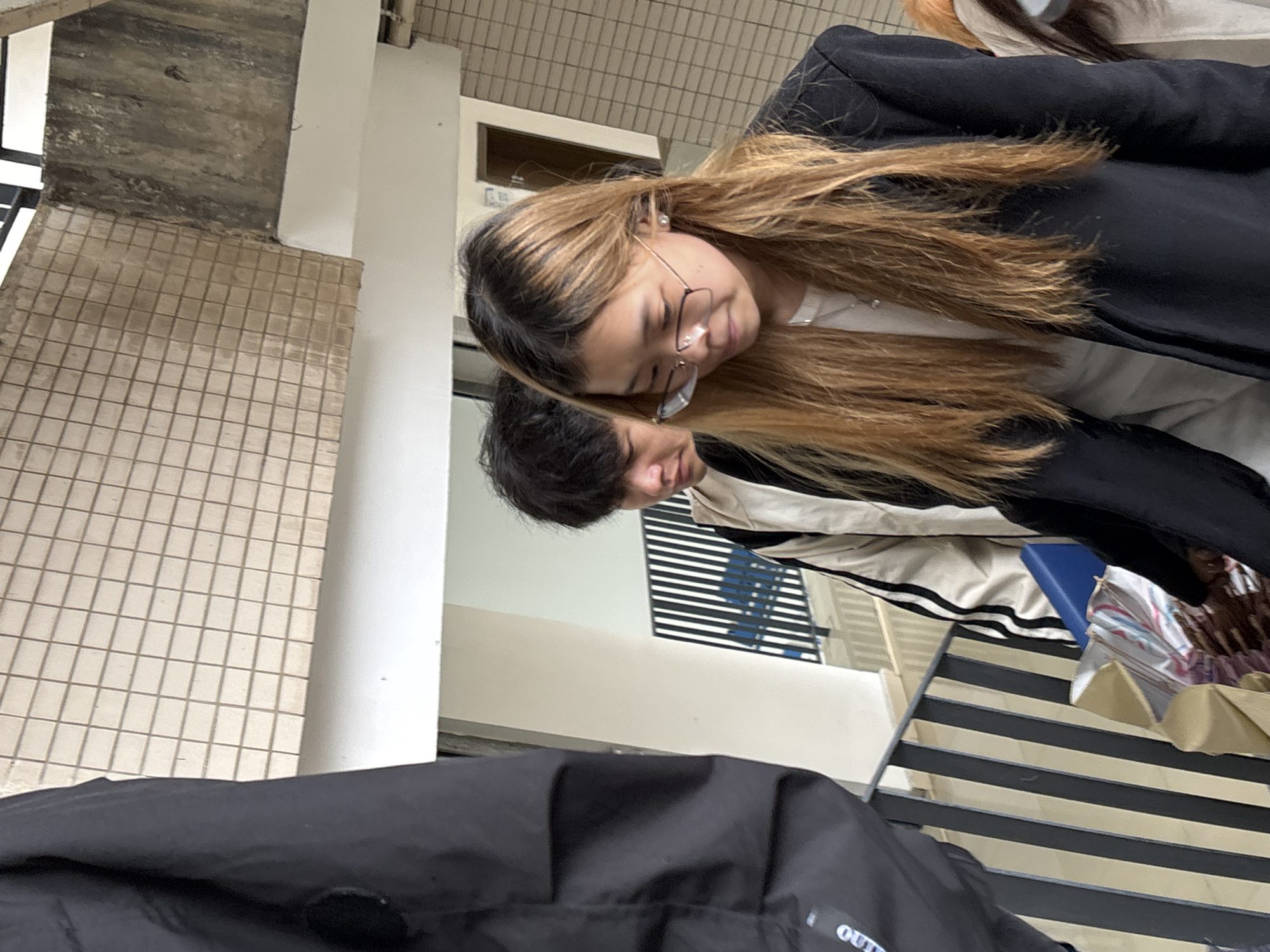 Two people are present in the scene: a woman with long, light brown hair and glasses, and a man with short, dark hair. They appear to be sitting close to each other. The woman is wearing a black blazer over a white top. The man is wearing a beige and black striped jacket. A black jacket obscures part of the lower foreground. The setting includes a tiled floor, a white pillar, and a structure with black and white stripes. A glimpse of a window with horizontal bars is visible in the background. There is insufficient information to identify the location in Jiangjin, China.