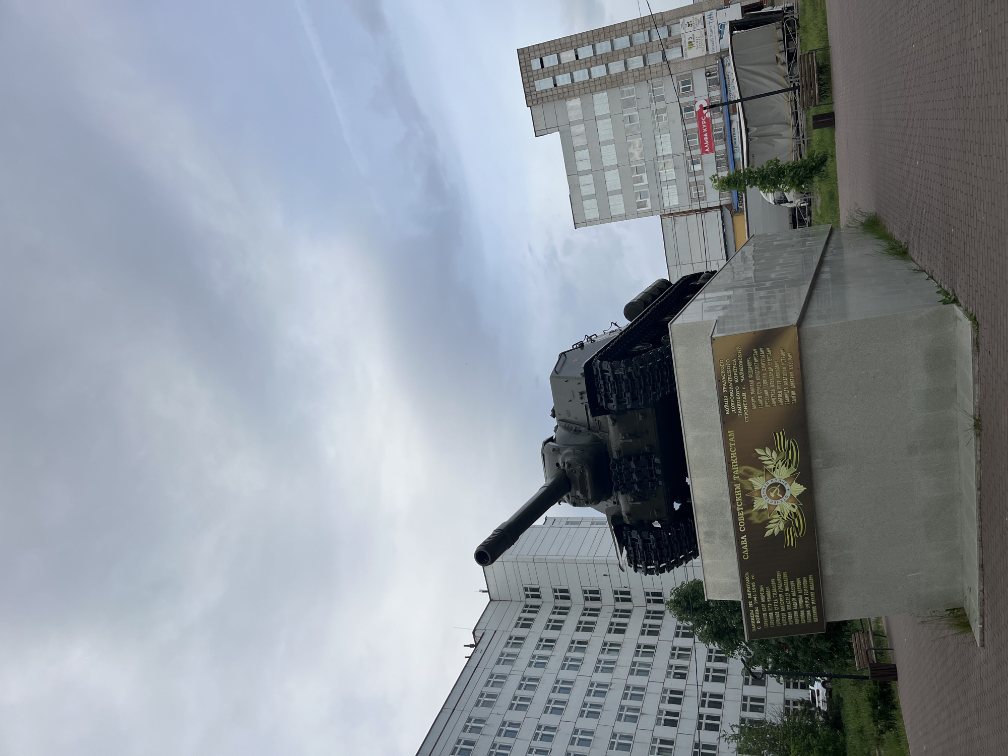 The image depicts a monument in the city of Chaikovsky, Russia, dedicated to Soviet tankists. The monument features a T-34 tank mounted on a concrete plinth. A plaque on the plinth is inscribed in Russian, honoring the tankists of the Ural Volunteer Tank Corps and builders of Chaikovsky. The inscription also lists names of local soldiers who did not return from World War II. The tank faces left in the image, with the gun barrel pointing towards the sky.
In the background, there are large apartment buildings. The sky is overcast. A red advertising banner is also partly visible in the background. The monument is surrounded by a paved area.